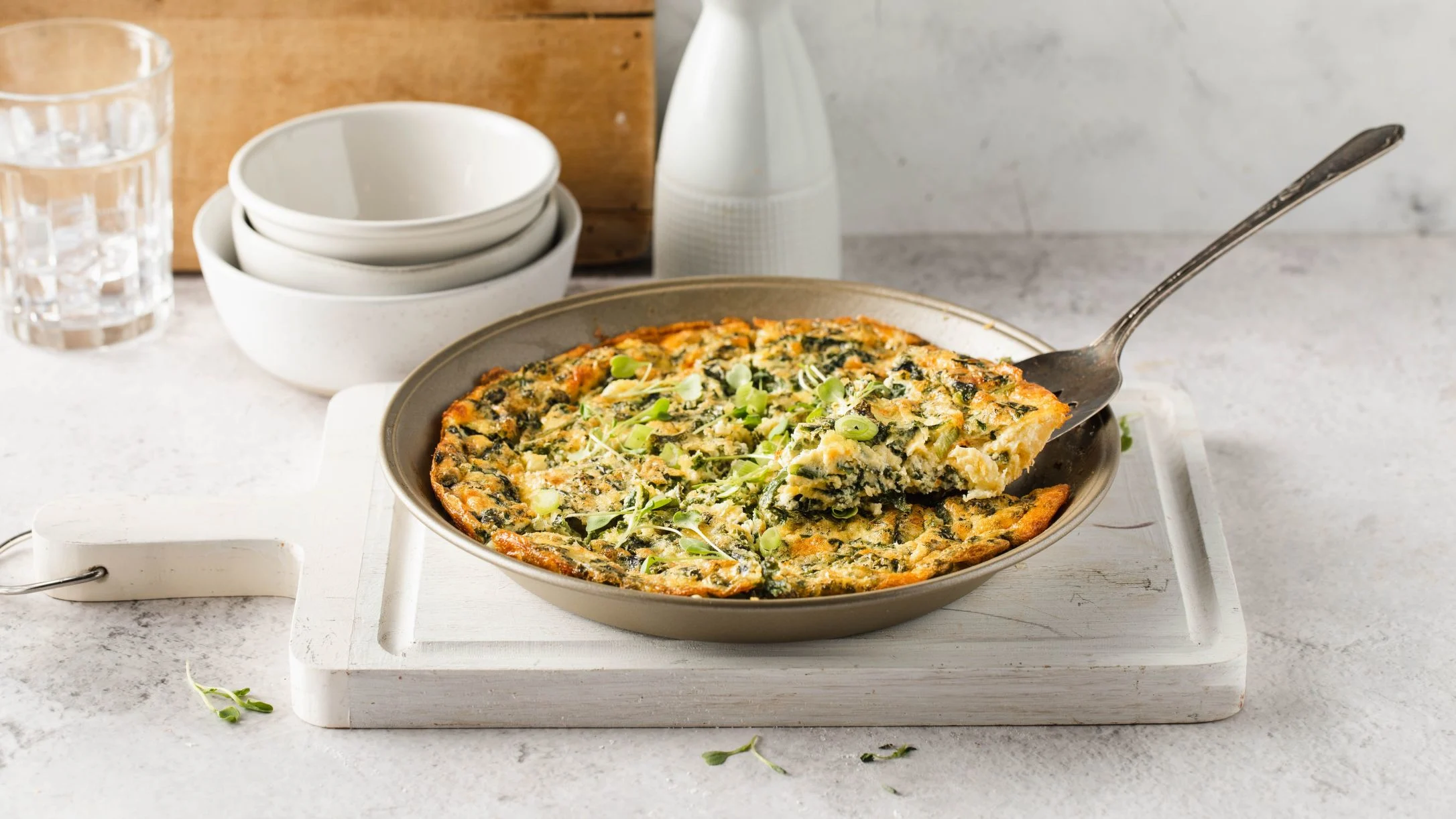 Spinach and herb frittata in a pan with serving spoon, stacked white bowls and water glass in background.