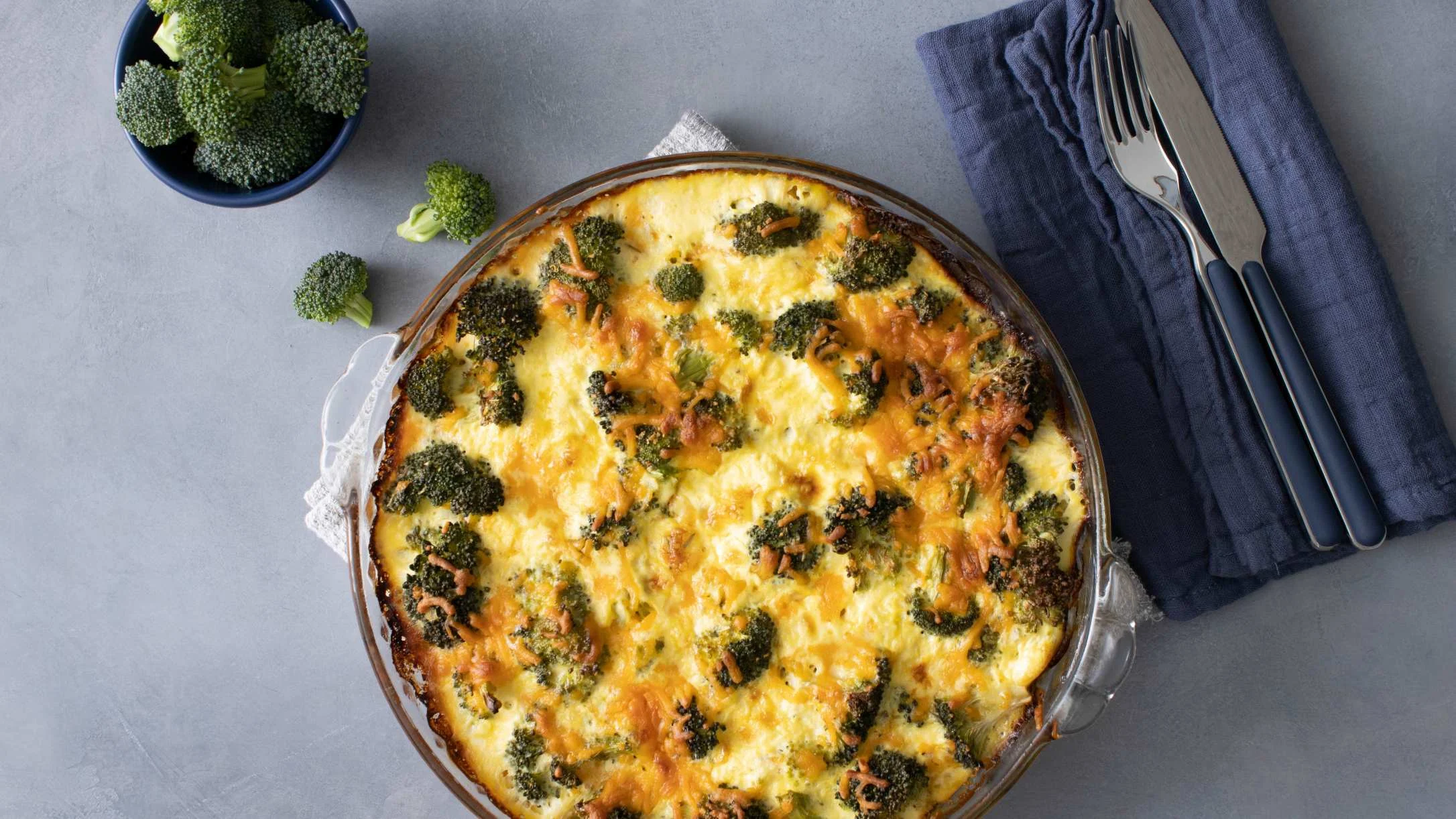 Cheesy broccoli quiche in glass dish with fresh broccoli in blue bowl and utensils on blue napkin.