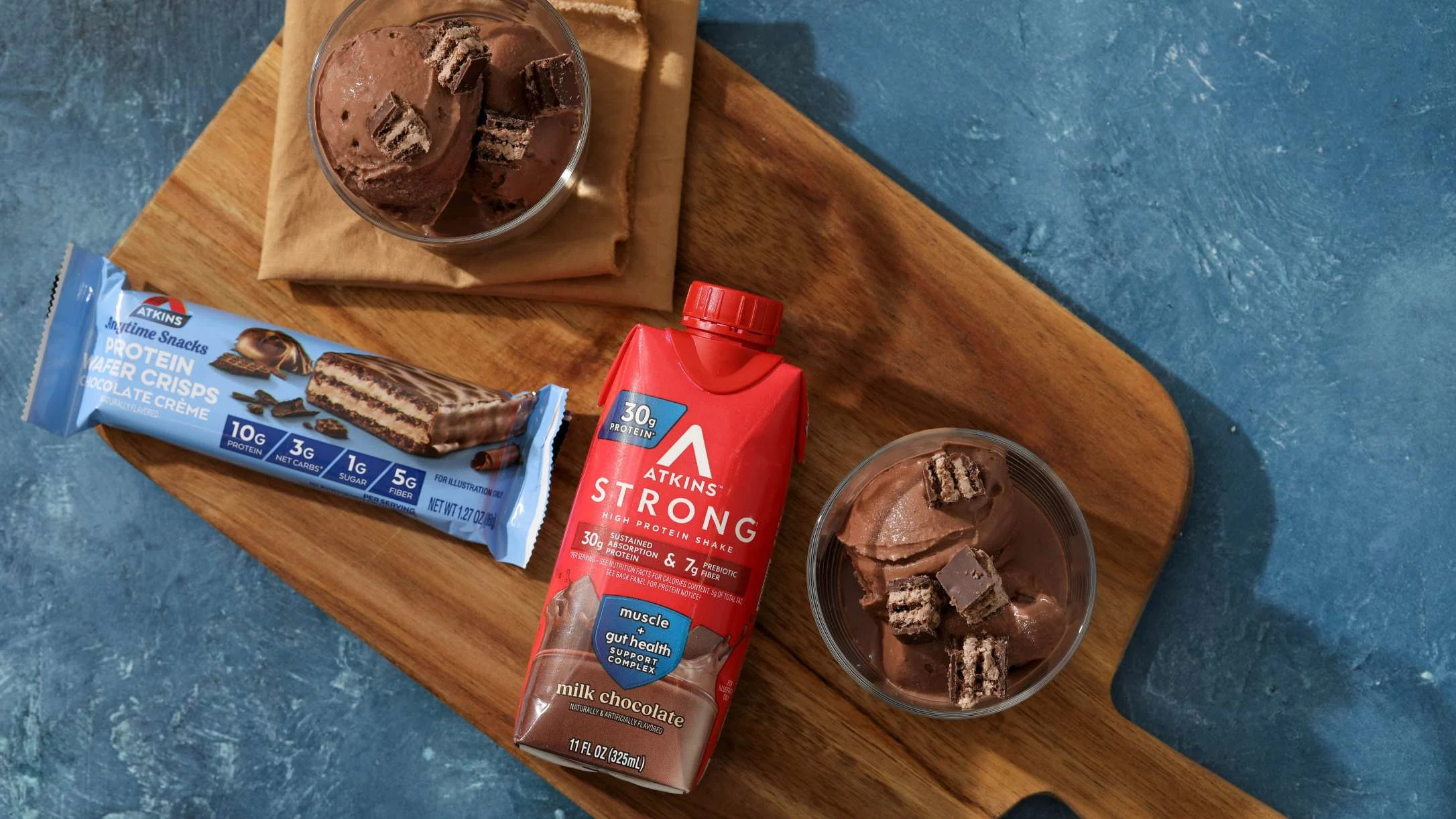 Atkins protein products on wooden board - chocolate shake, protein bar, and chocolate ice cream with cookie pieces.