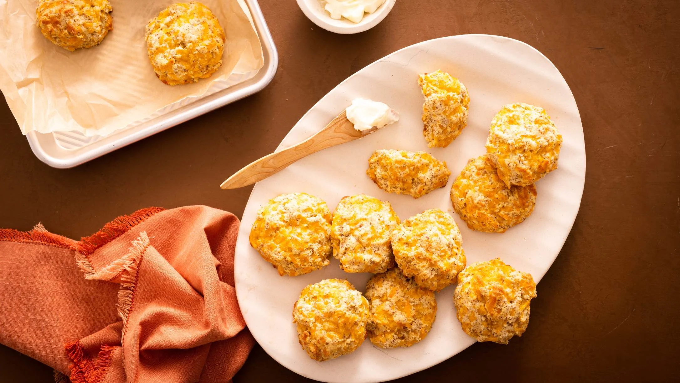Golden cheese biscuits on white oval platter with wooden butter knife, baking tray, and orange napkin on brown table.