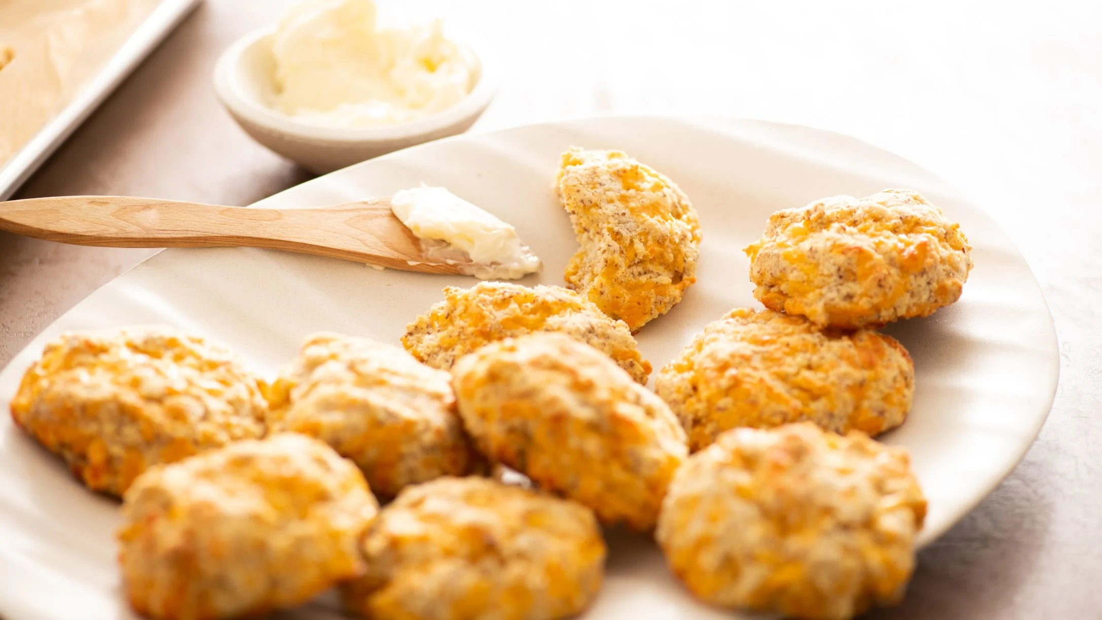 Freshly baked cheddar biscuits on a white plate with a wooden spoon of butter and a small bowl of butter nearby.