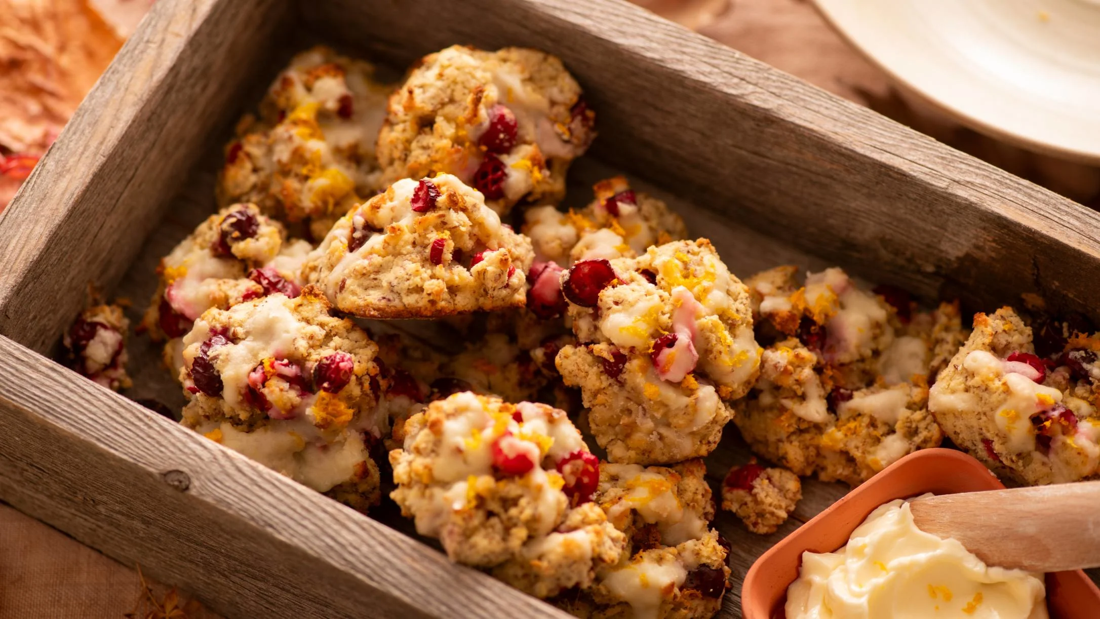 Freshly baked cranberry orange scones with glaze in a wooden box, with a small dish of butter visible.