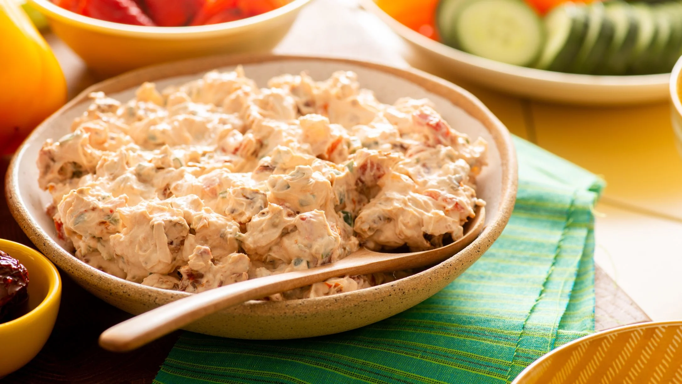 Creamy dip in a beige bowl with wooden spoon, surrounded by vegetable platters on a green placemat.
