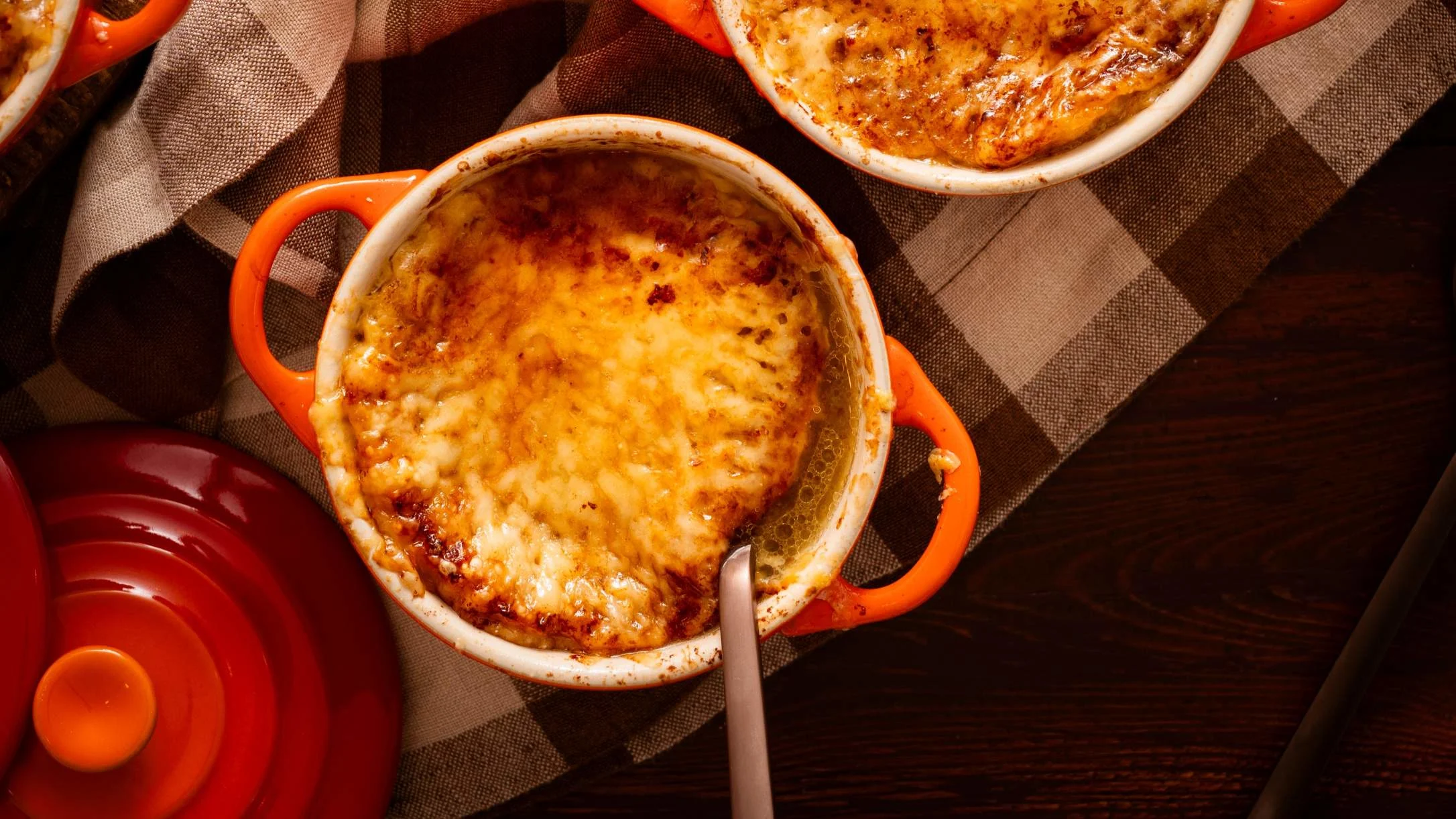 Bubbling cheese-topped French onion soup in bright orange ramekins on a checkered cloth, with a spoon ready for serving.