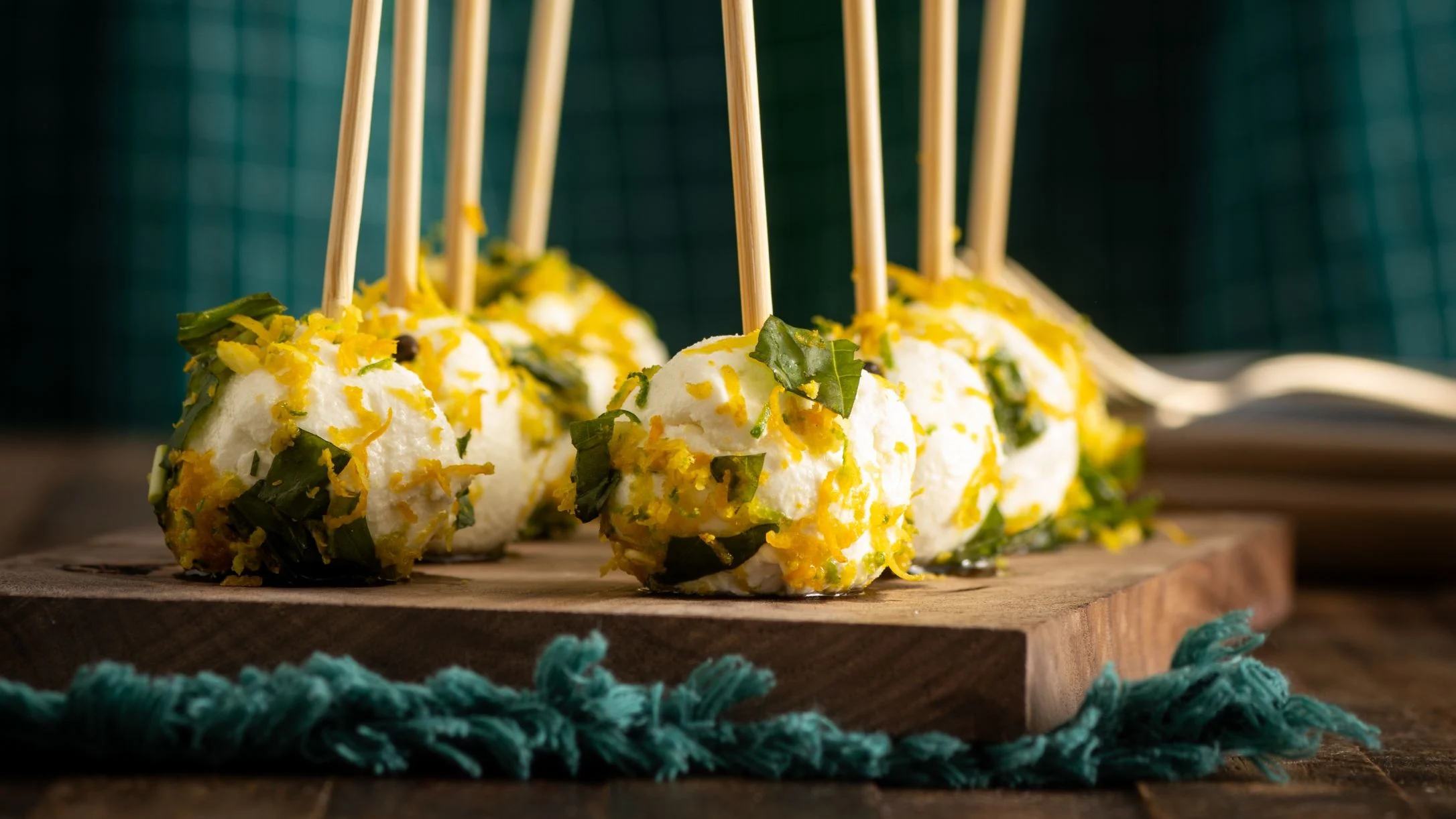 Cheese appetizers on wooden sticks garnished with herbs and citrus zest, displayed on a wooden serving board.