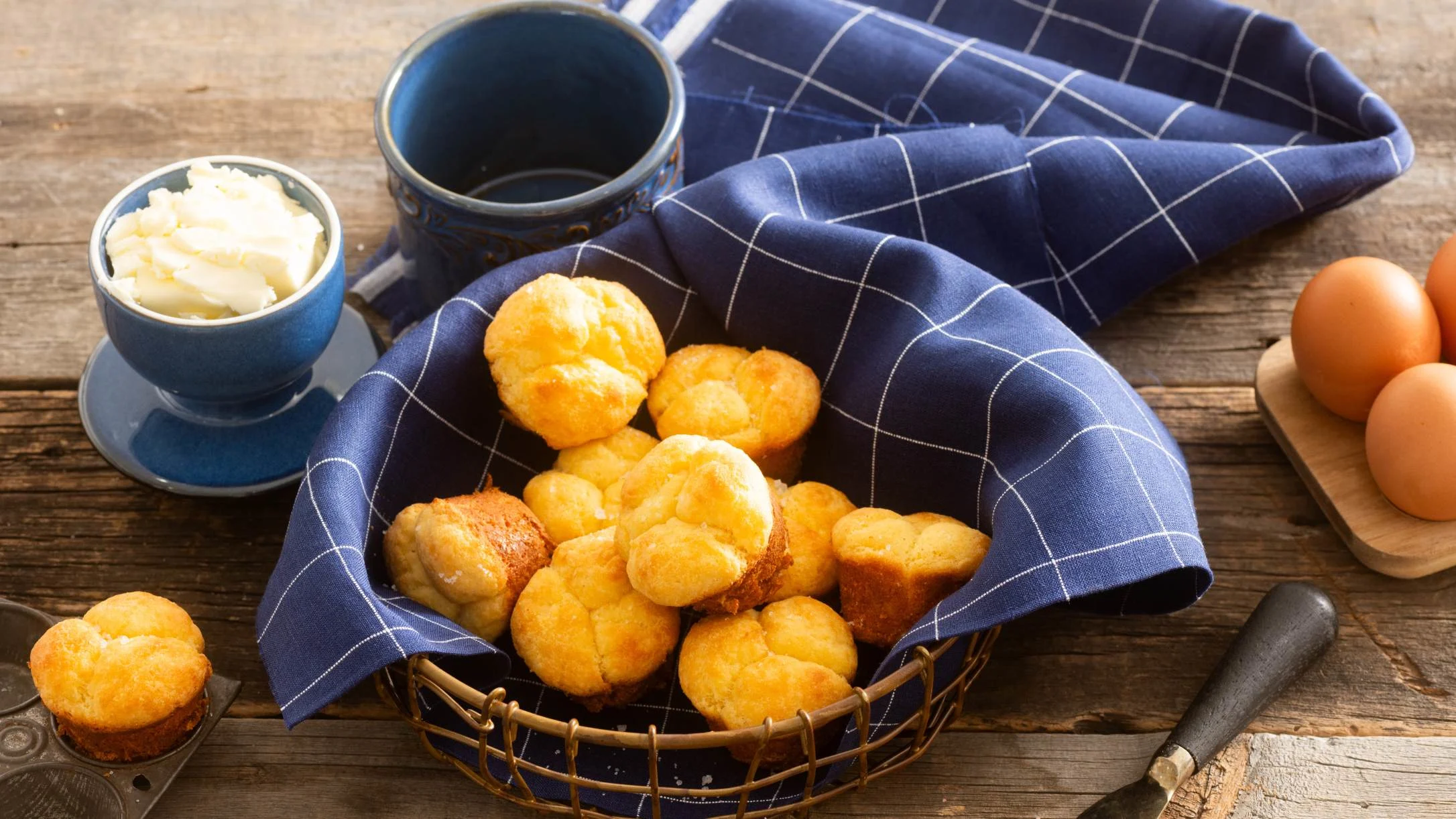 Golden keto rolls in basket with blue napkin, butter dish, coffee mug, and eggs on wooden table.