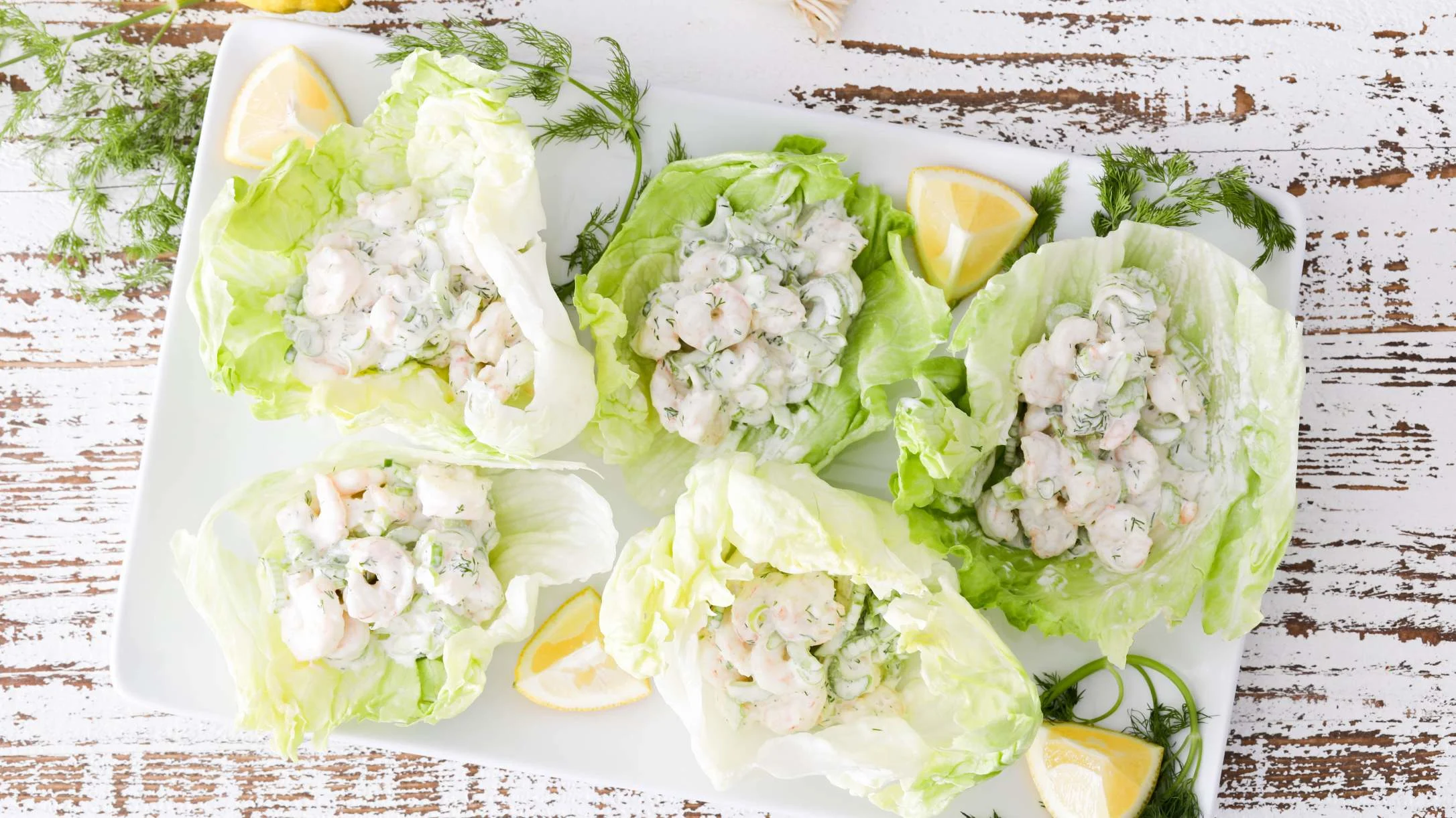 Shrimp salad lettuce cups with creamy dill dressing and lemon wedges served on a white platter on rustic wooden surface.