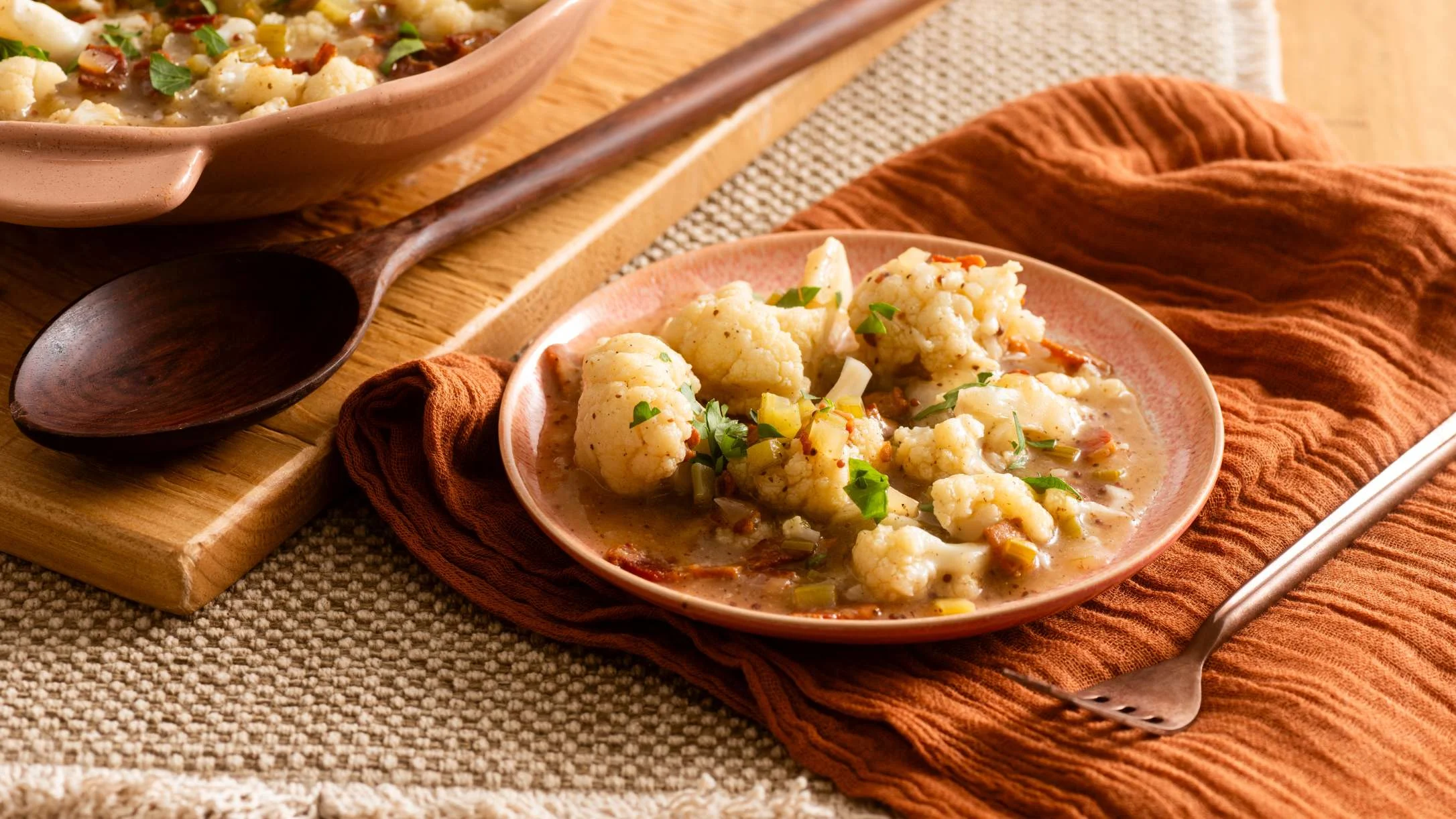 Cauliflower with bacon and herbs served in terracotta dishes on wooden board with rustic orange napkin and wooden spoon.