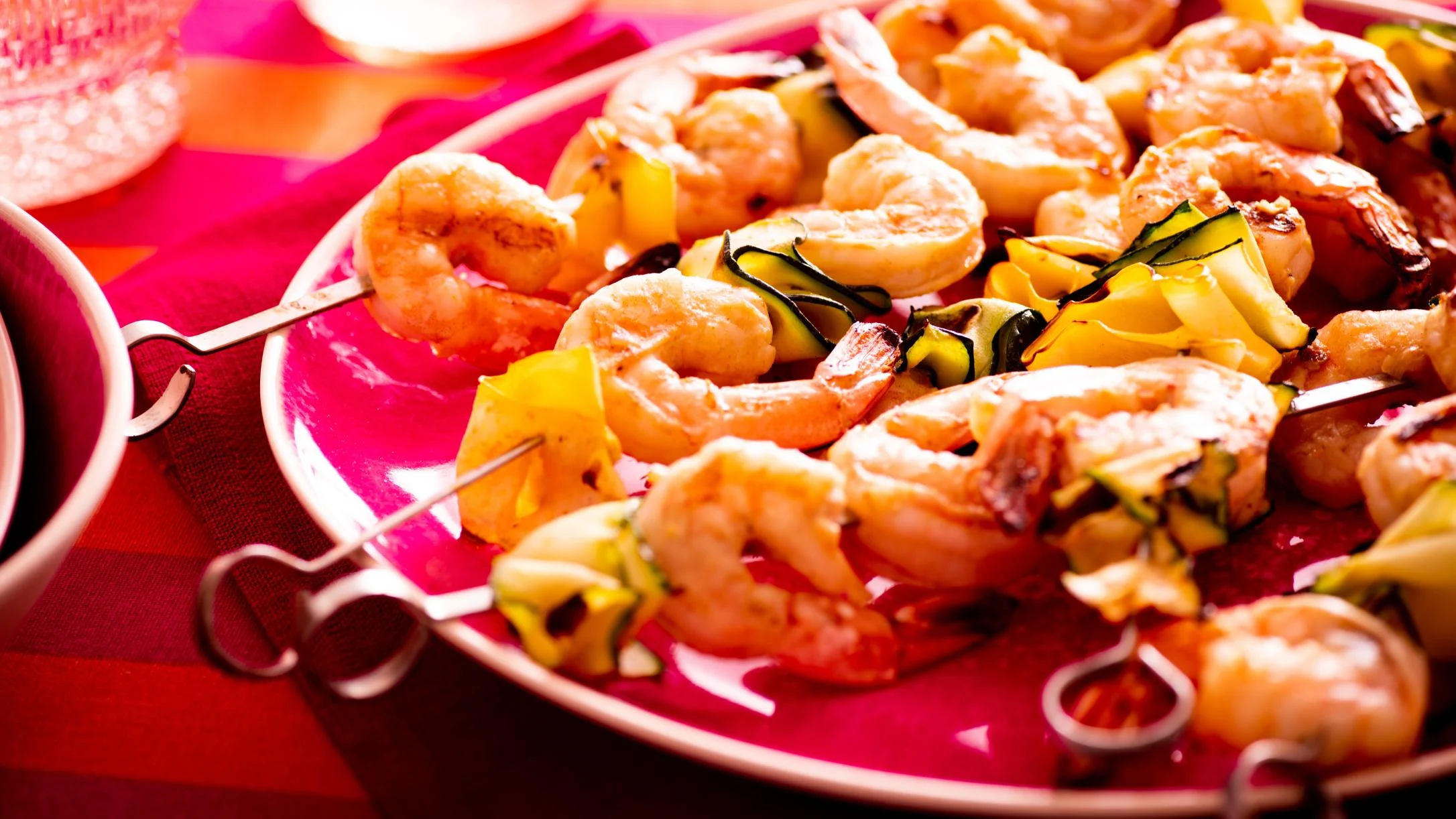 Grilled shrimp skewers with yellow and green vegetables served on a bright pink plate against a red background.