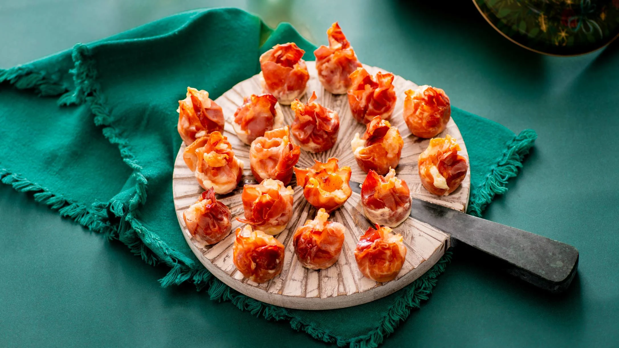 Crispy prosciutto-wrapped appetizers arranged on a wooden serving board with a knife, placed on teal fabric.