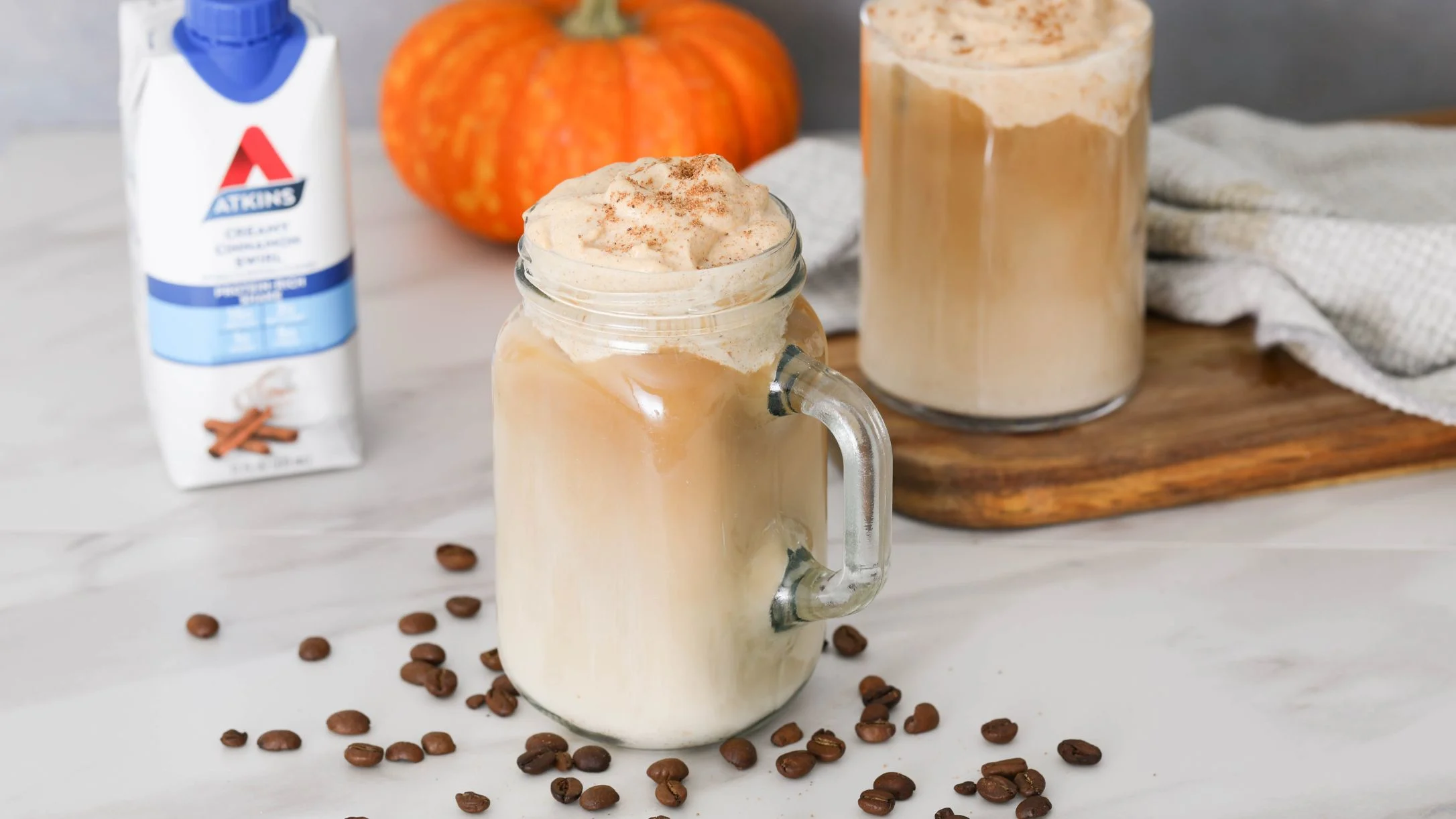 Pumpkin spice lattes in glass mugs with whipped cream, surrounded by coffee beans, Atkins protein shake, and a small pumpkin.