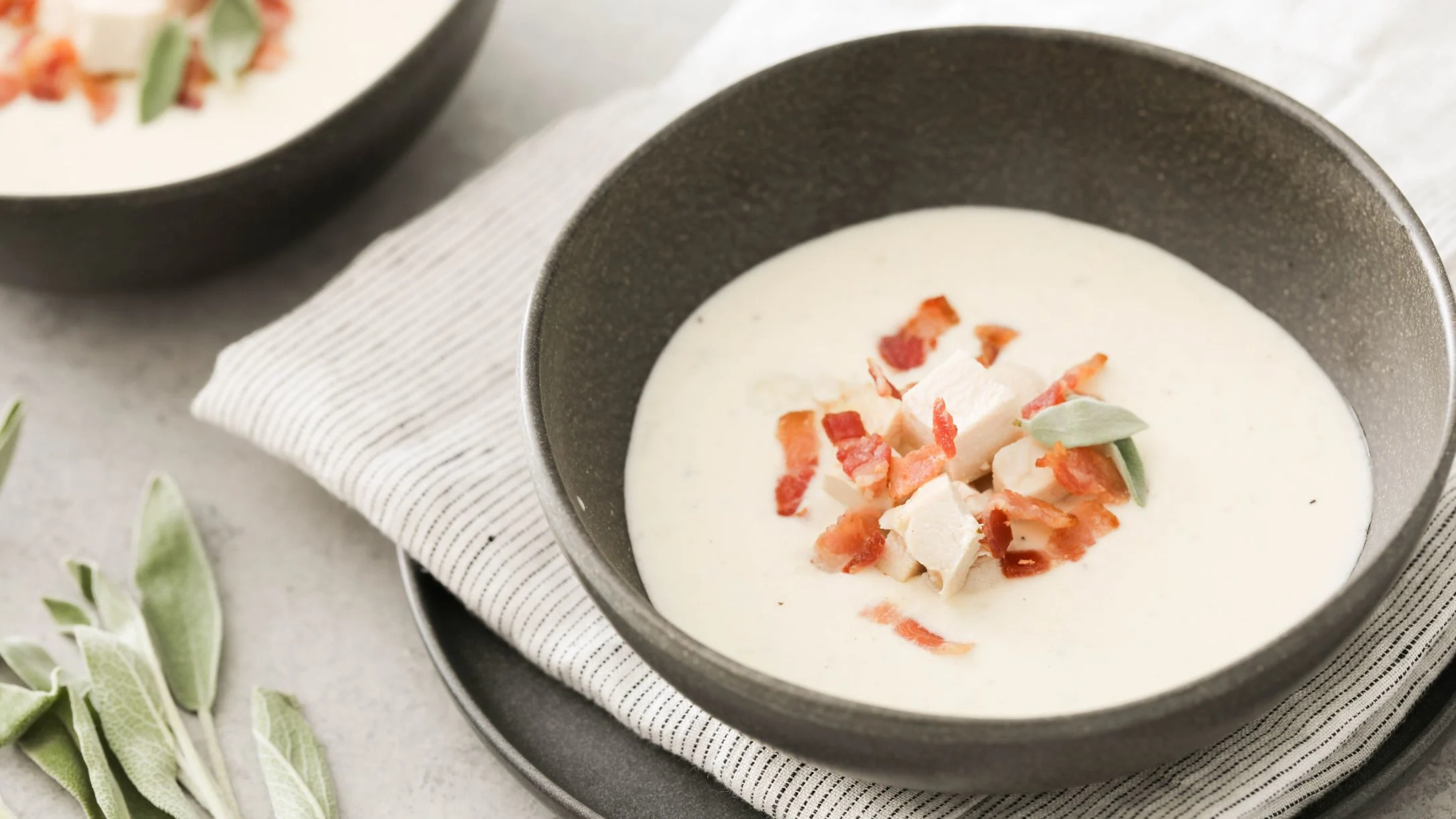 Creamy white soup in dark bowl topped with diced bacon and chicken, garnished with sage leaves on striped cloth.
