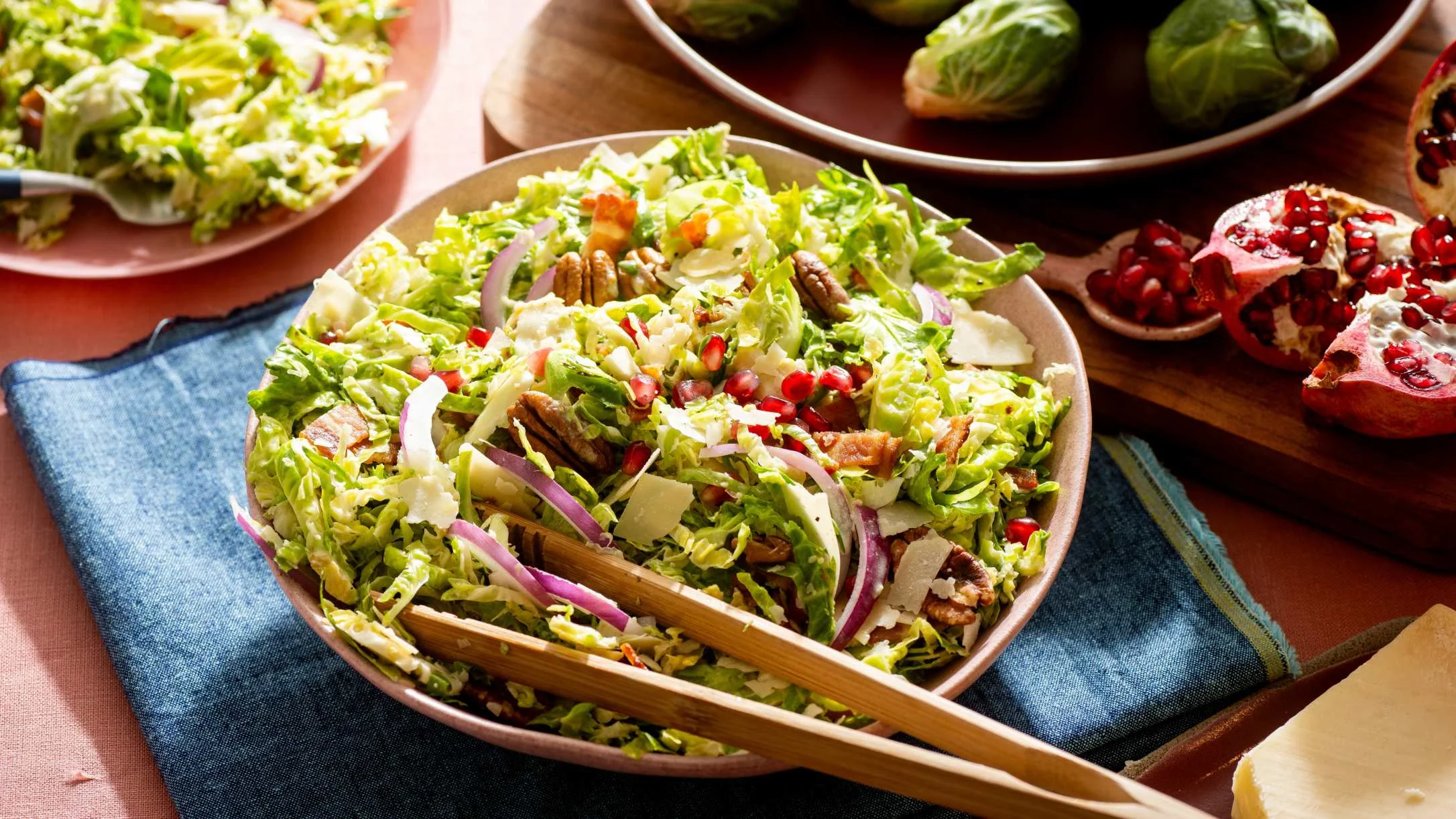 Fresh Brussels sprout salad with pecans, pomegranate seeds, and red onion in ceramic bowl with wooden serving utensils.