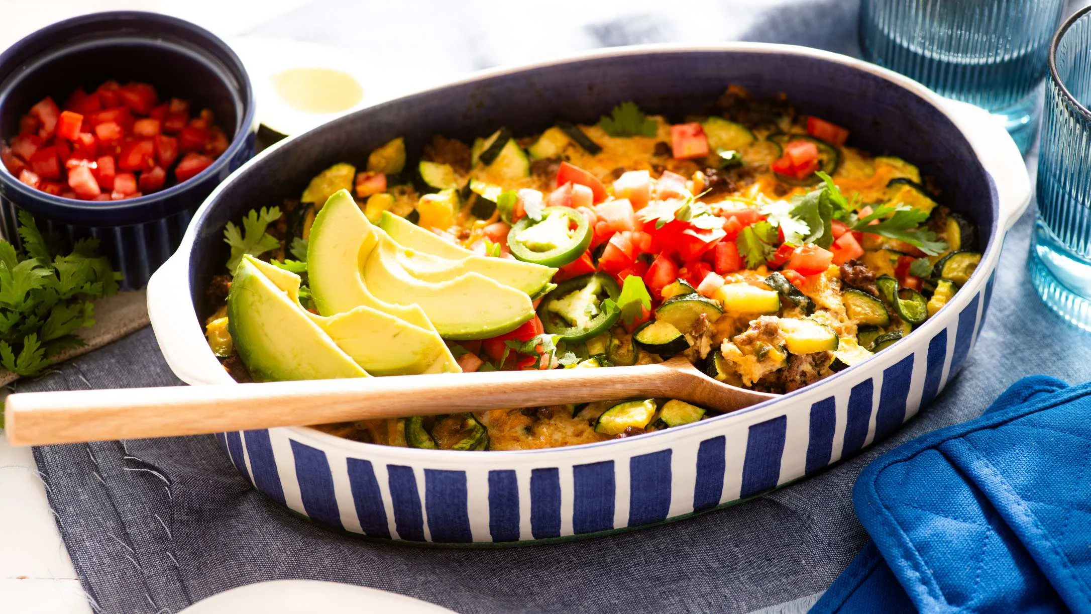 Mexican casserole in striped dish topped with avocado slices, diced tomatoes and cilantro, with salsa on the side.