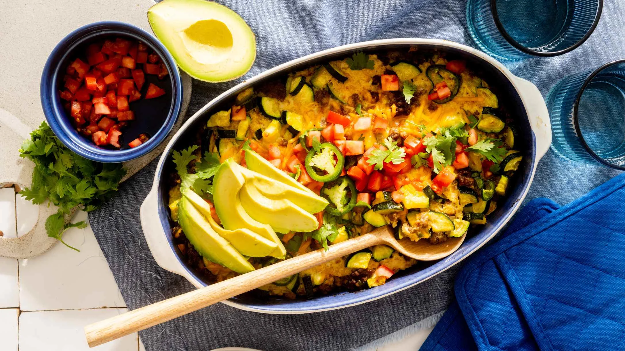 Mexican casserole topped with tomatoes, avocado slices, and cilantro, served with salsa in a blue bowl.