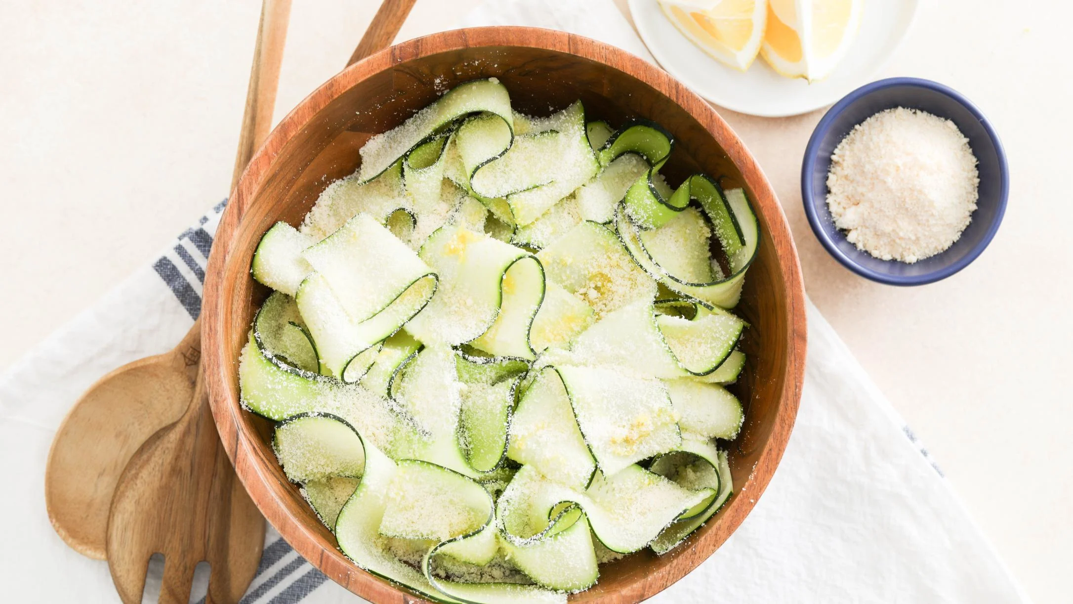 Zucchini ribbons with cheese and lemon zest in a wooden bowl with wooden utensils and ingredients surrounding it.