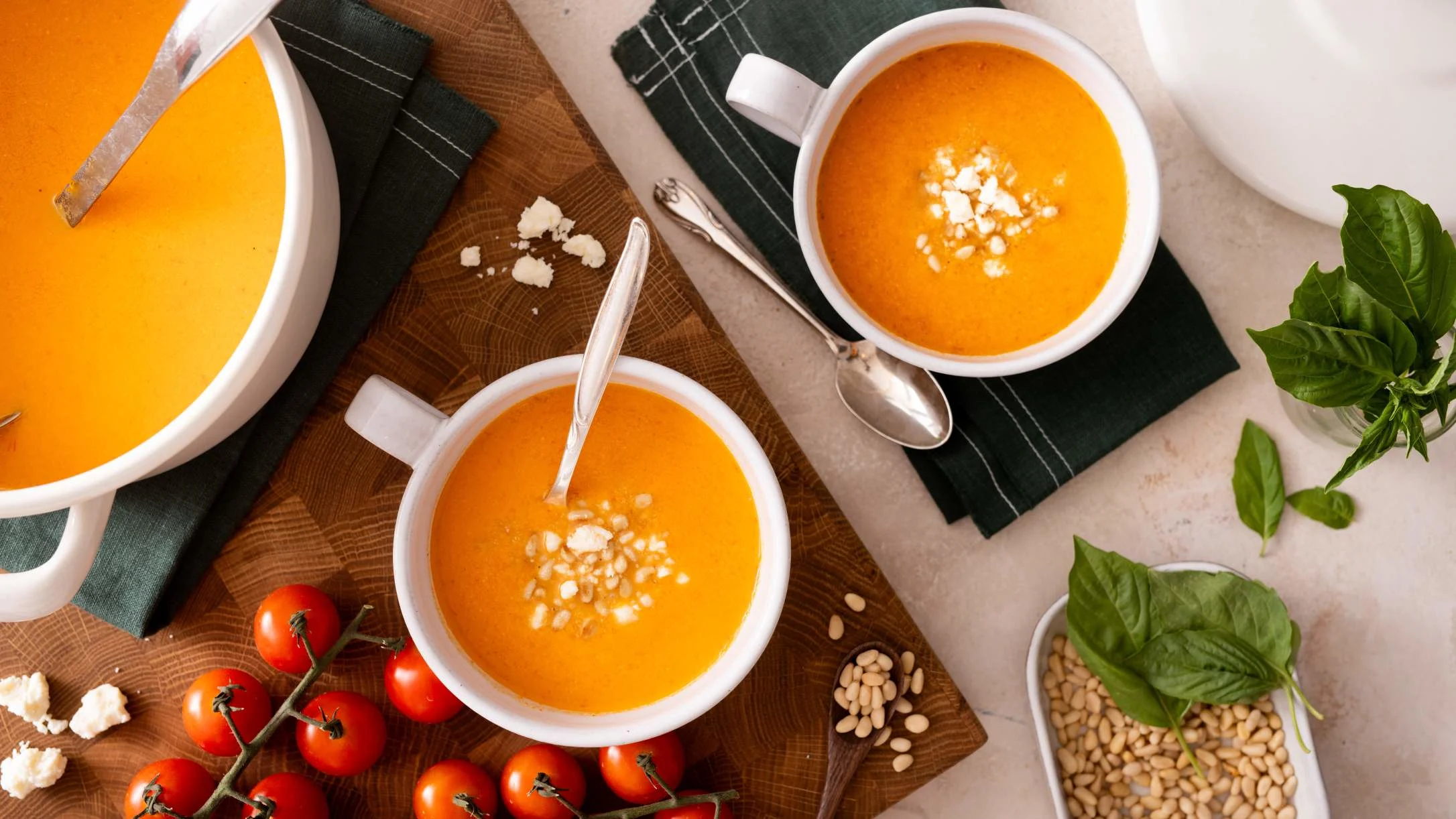 Bowls of orange tomato soup garnished with cheese, served with fresh tomatoes, basil, and pine nuts on wooden board.