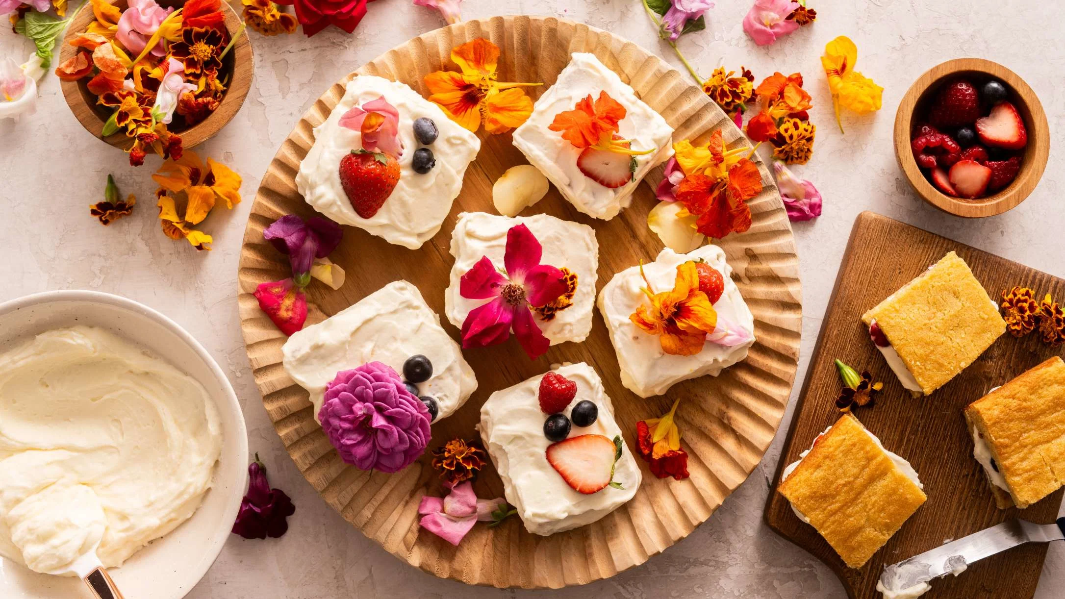 Mini cakes decorated with edible flowers and berries on a wooden platter, surrounded by frosting bowl and cake squares.