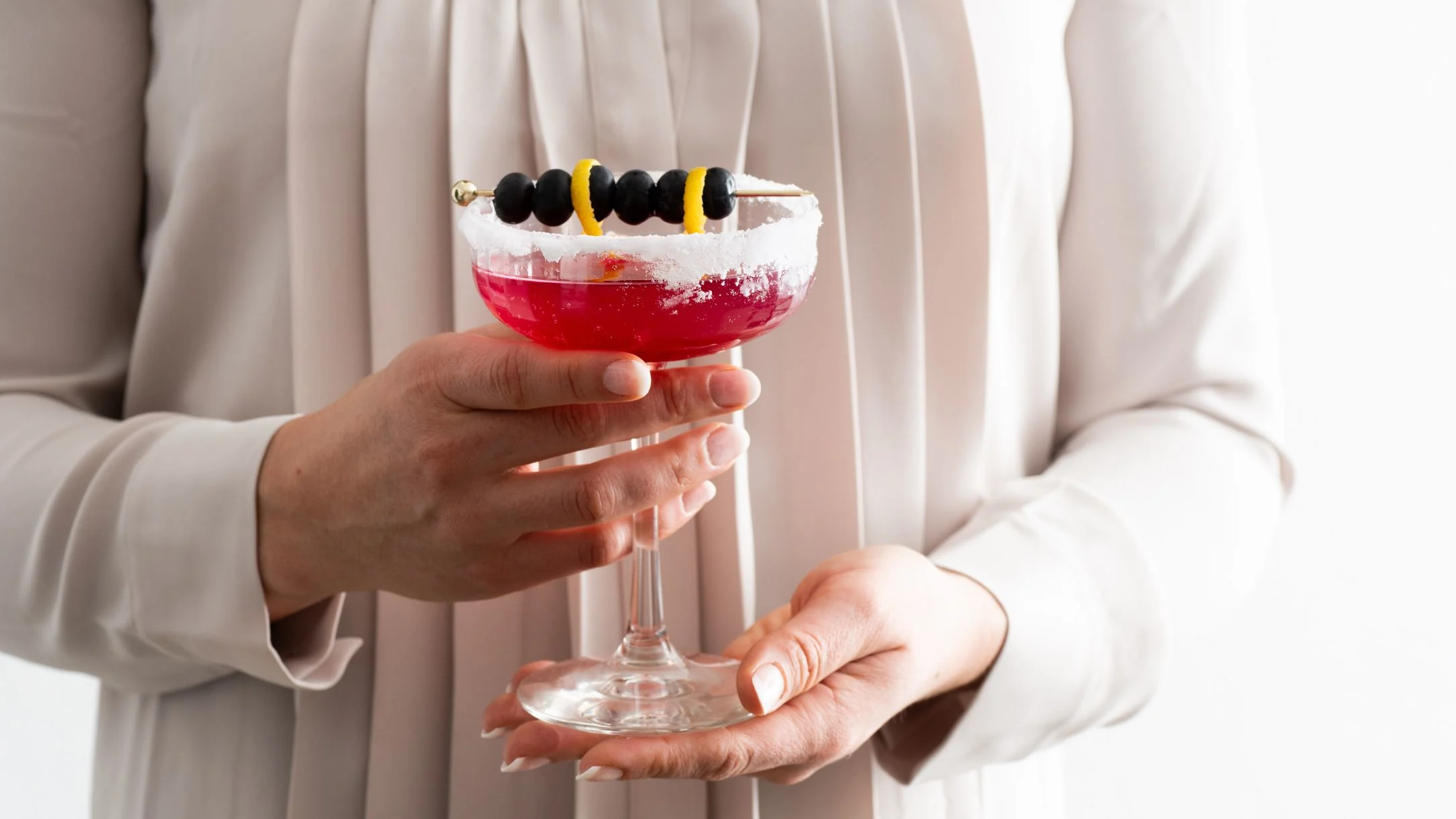 Person in white clothing holding a red cocktail with sugar-rimmed glass, garnished with blueberries and lemon twist.