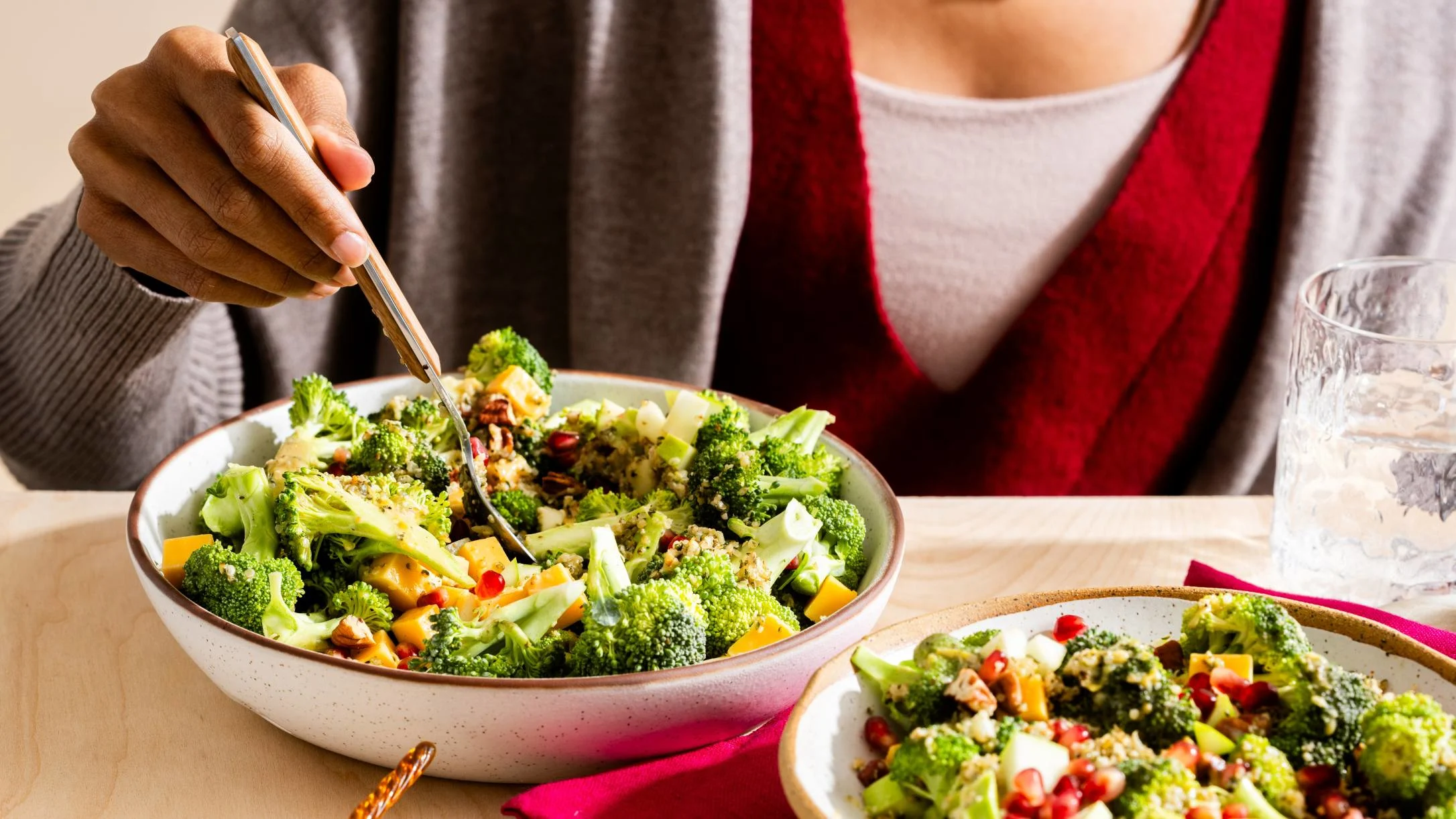 Person in red scarf eating a colorful broccoli salad with nuts and vegetables from a white bowl.