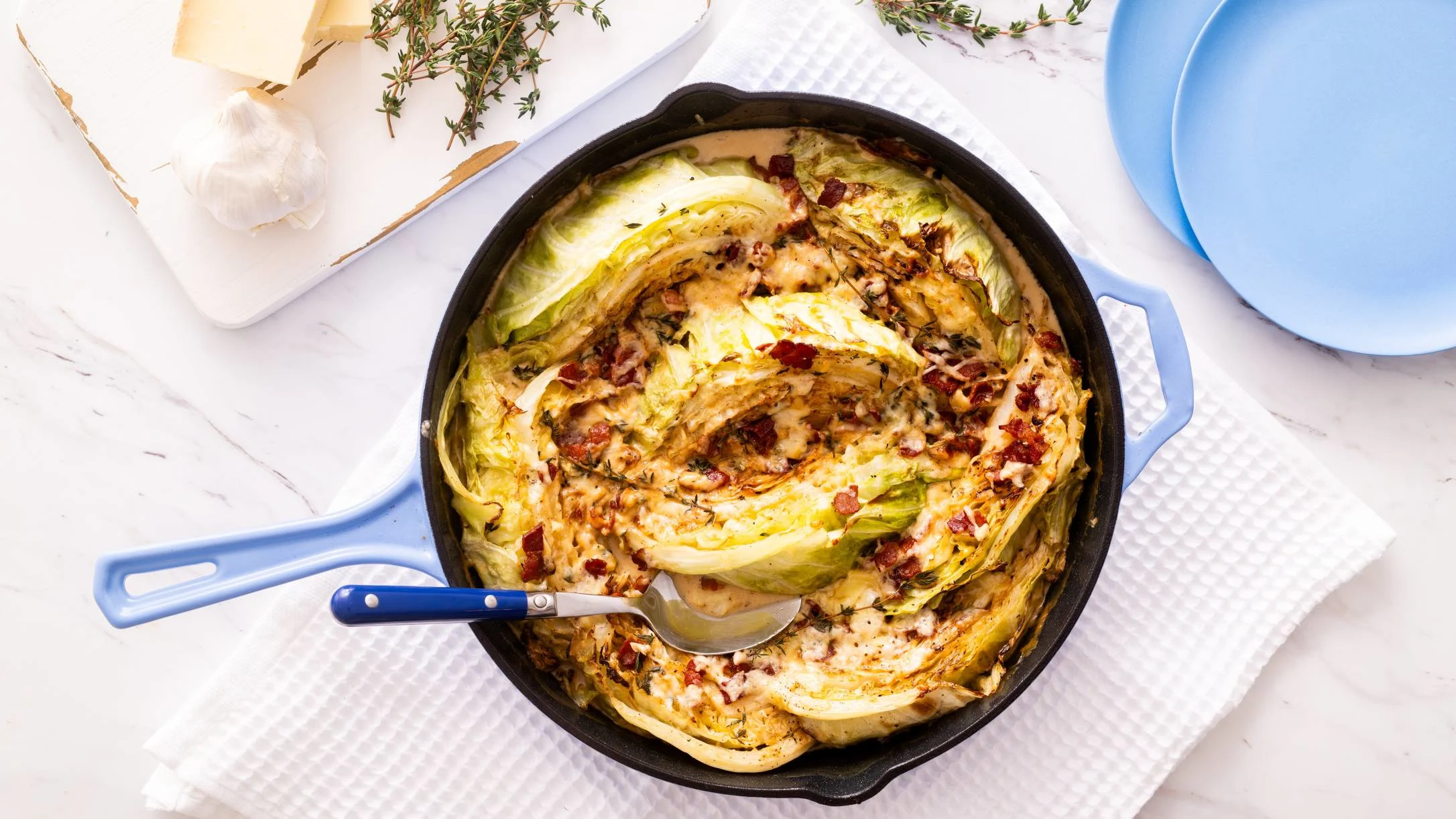 Roasted cabbage dish in blue cast iron skillet with cheese and bacon bits, served with fresh herbs and cheese nearby.