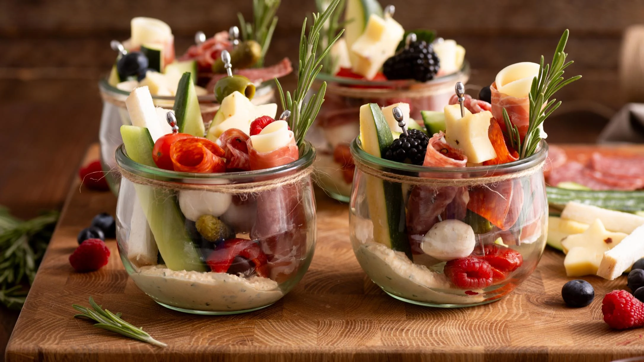 Glass jars filled with charcuterie appetizers featuring cheese, meats, berries, and vegetables garnished with rosemary sprigs.