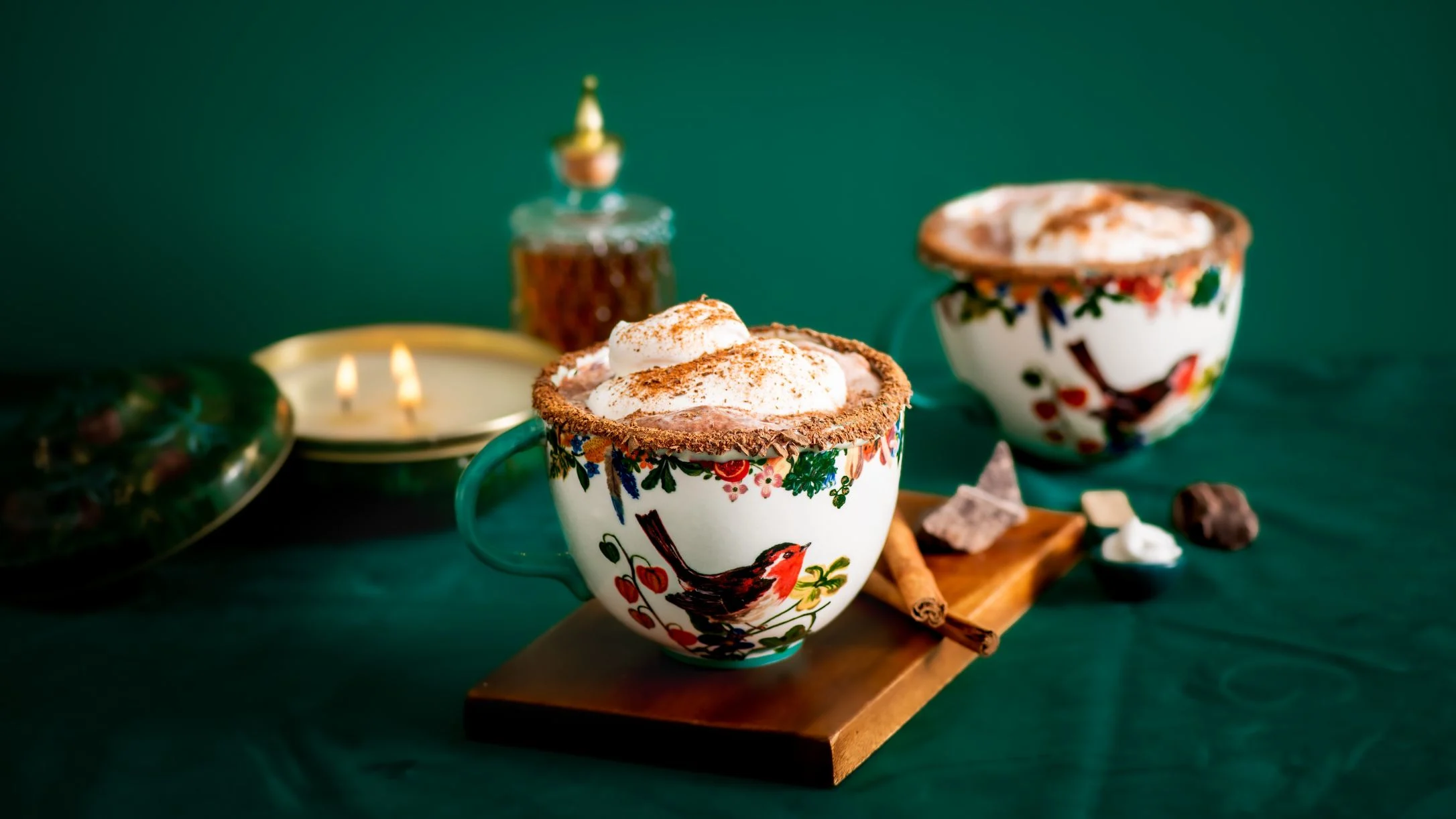 Festive mugs with hot chocolate topped with whipped cream and cinnamon, decorated with robin designs against a green background.
