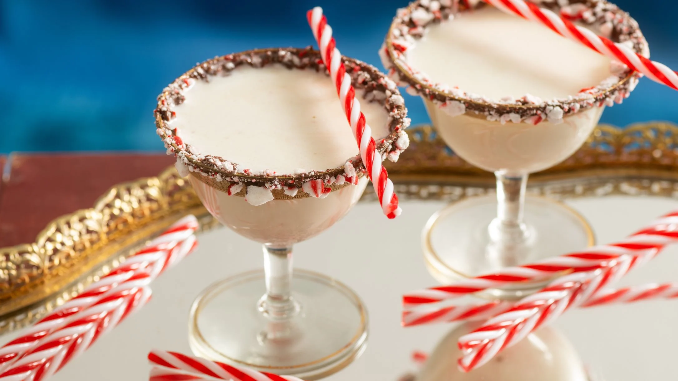Festive cocktails with peppermint candy cane rims and striped straws on an elegant gold tray against a blue background.