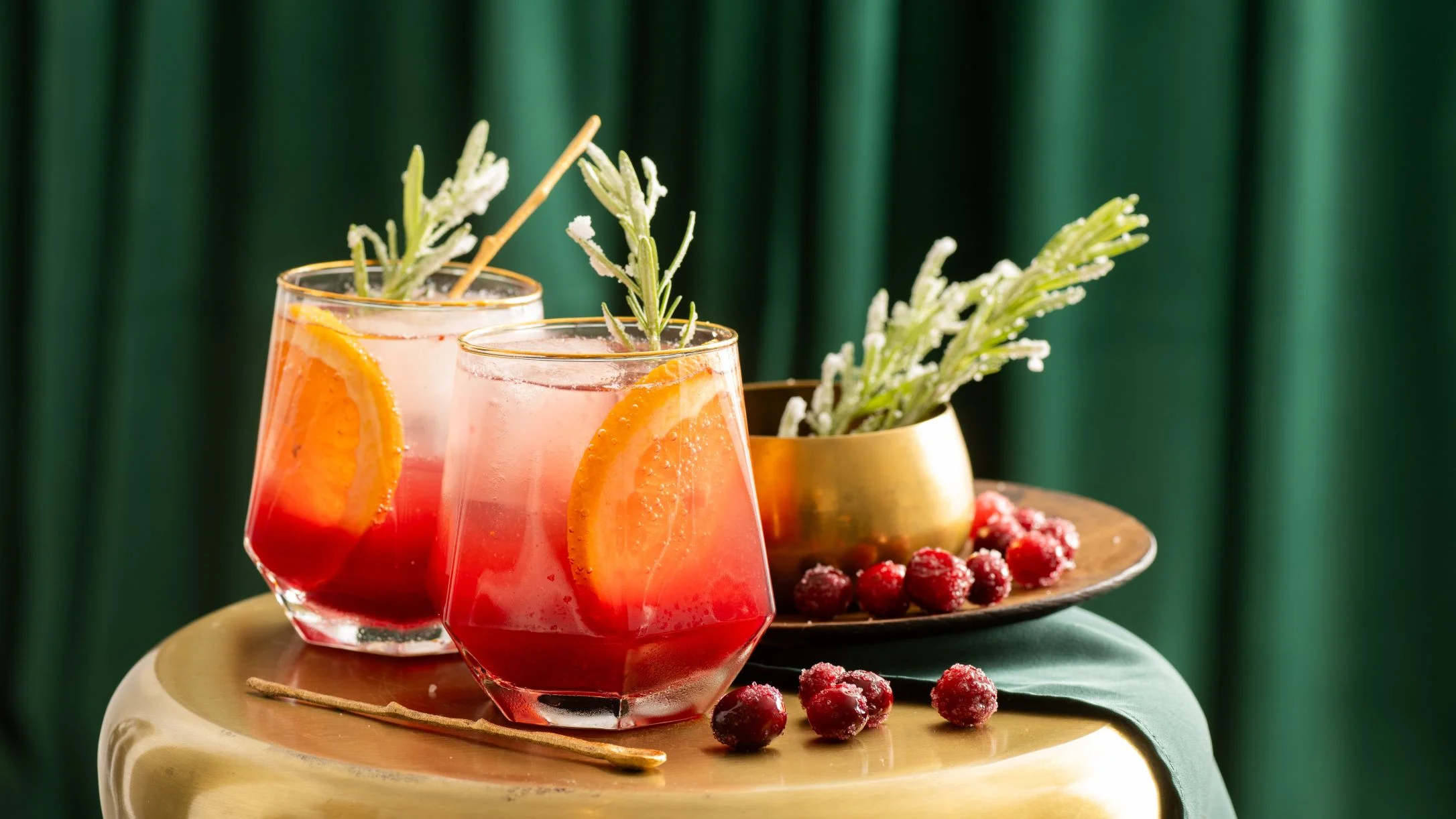 Two red cocktails with orange slices and rosemary garnish on gold tray with cranberries against green backdrop.