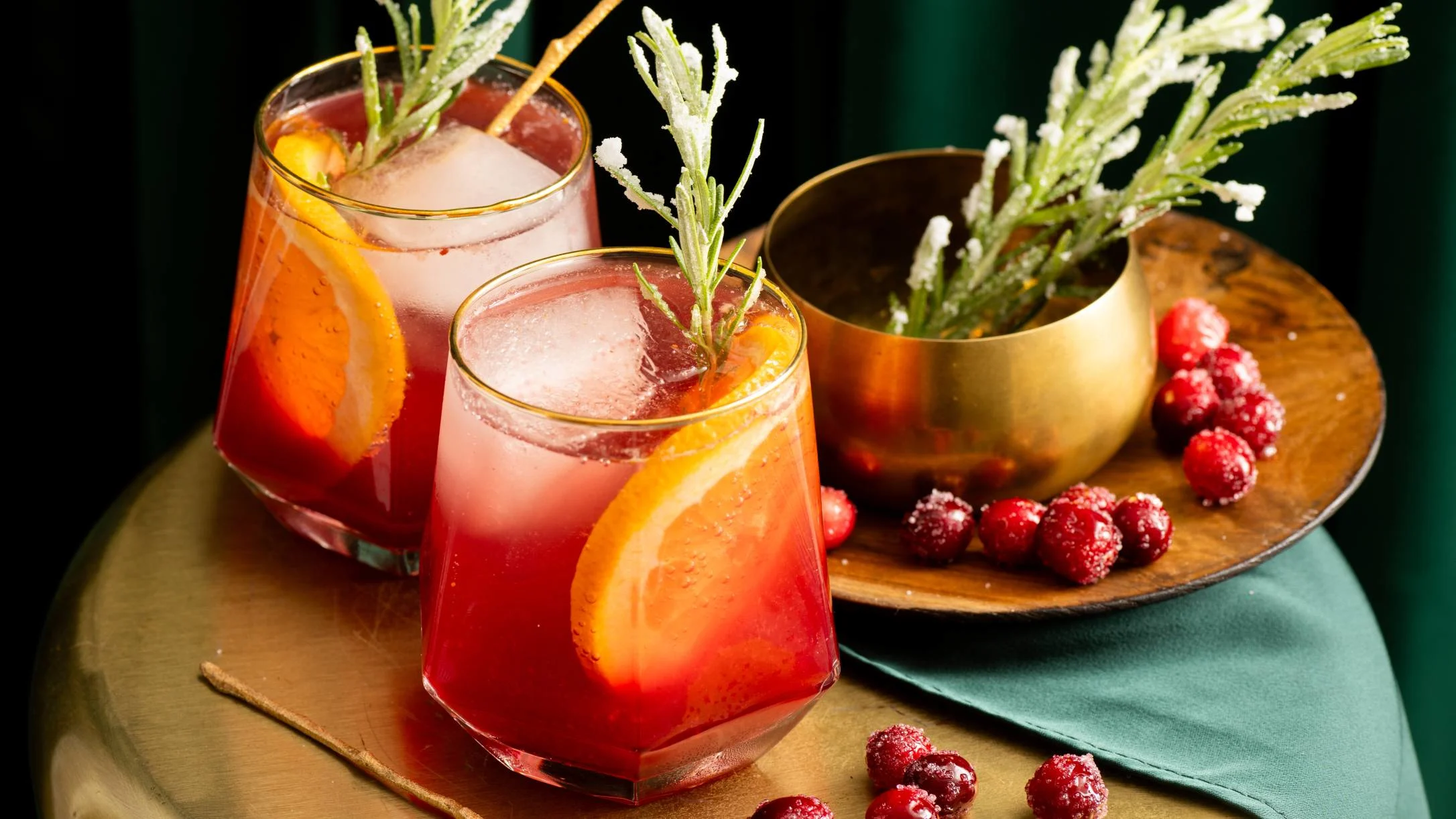 Two red cocktails with orange slices and rosemary garnish, served with frosted cranberries and a gold herb container.