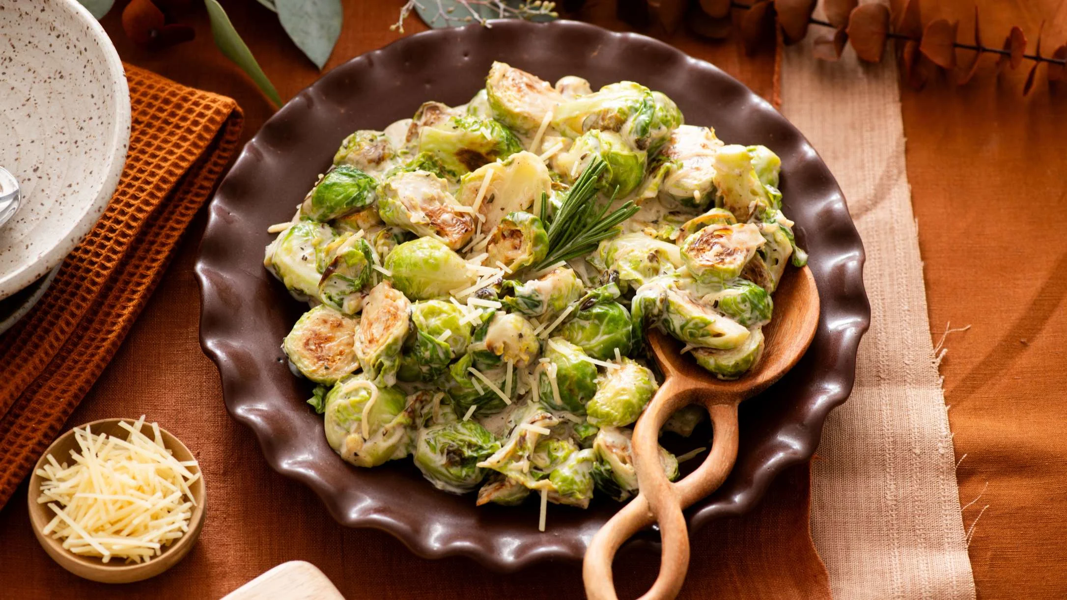 Creamy Brussels sprouts with cheese in a brown ceramic dish with wooden serving utensils on a rustic table.