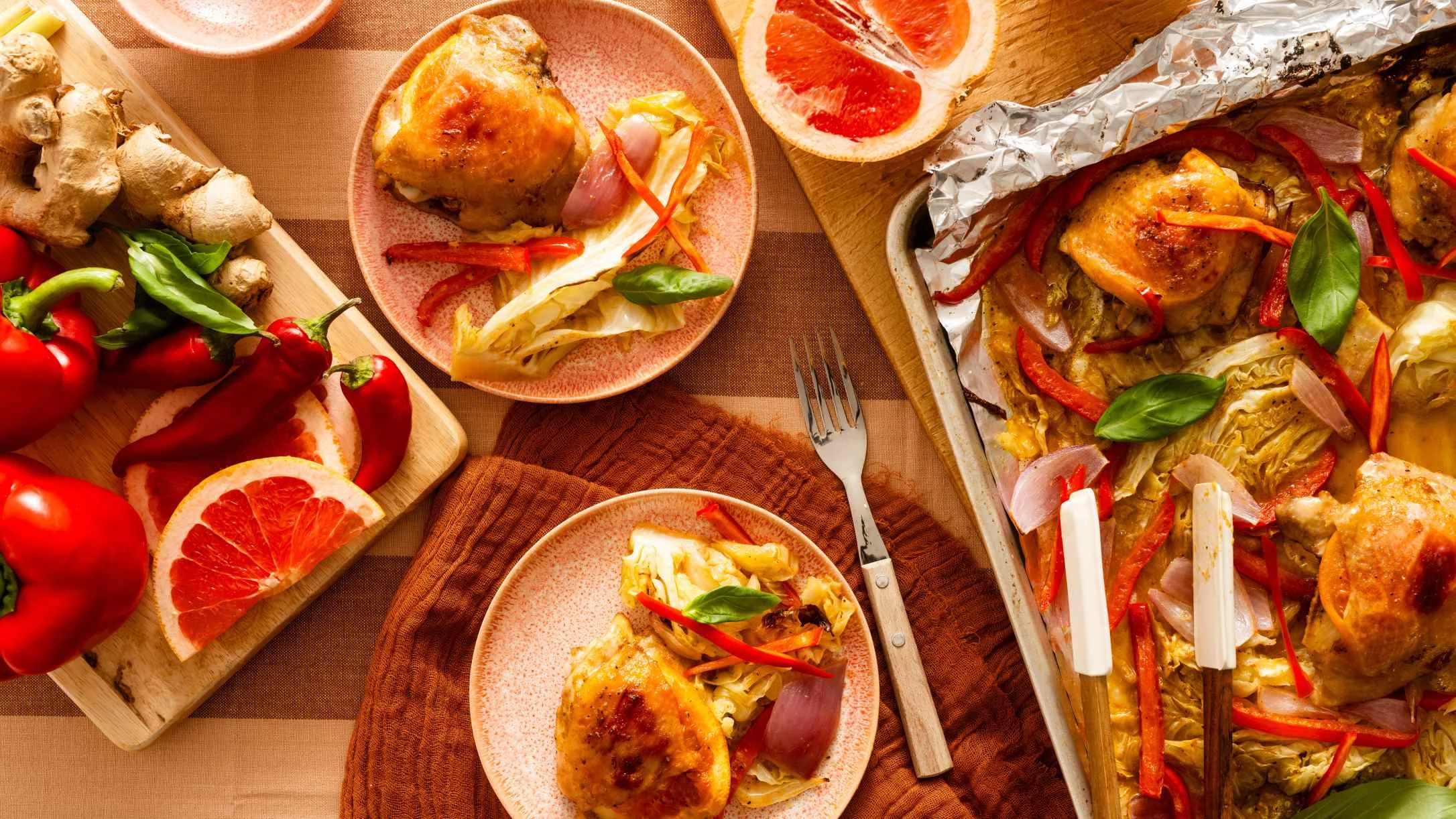 Roasted chicken dinner with cabbage, red peppers, and grapefruit slices on pink plates with fresh ingredients nearby.