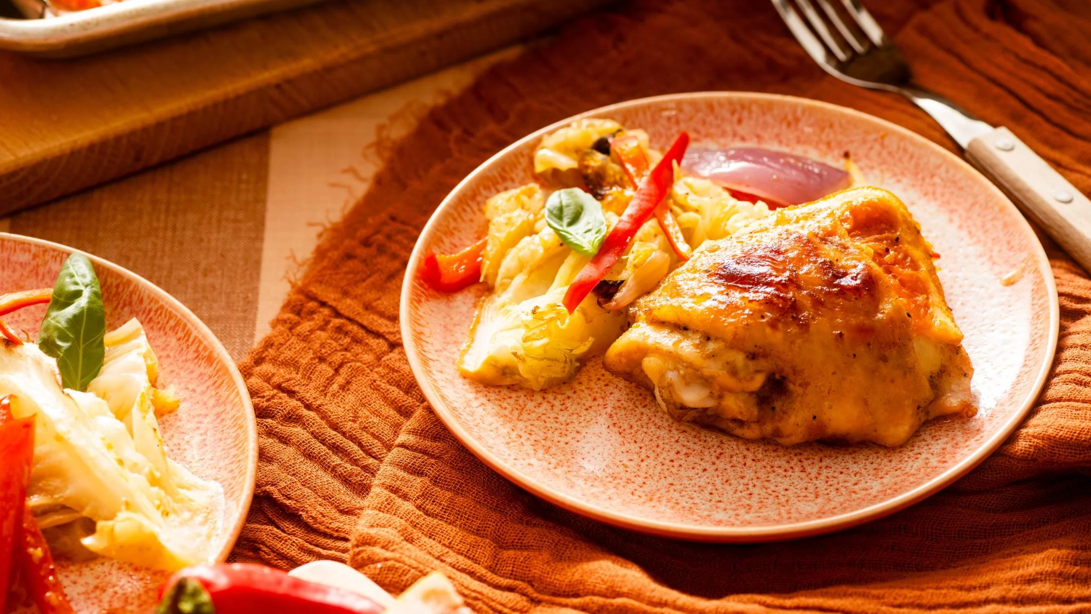 Roasted chicken thigh with vegetables on pink speckled plates, garnished with basil and red peppers on a wooden table.