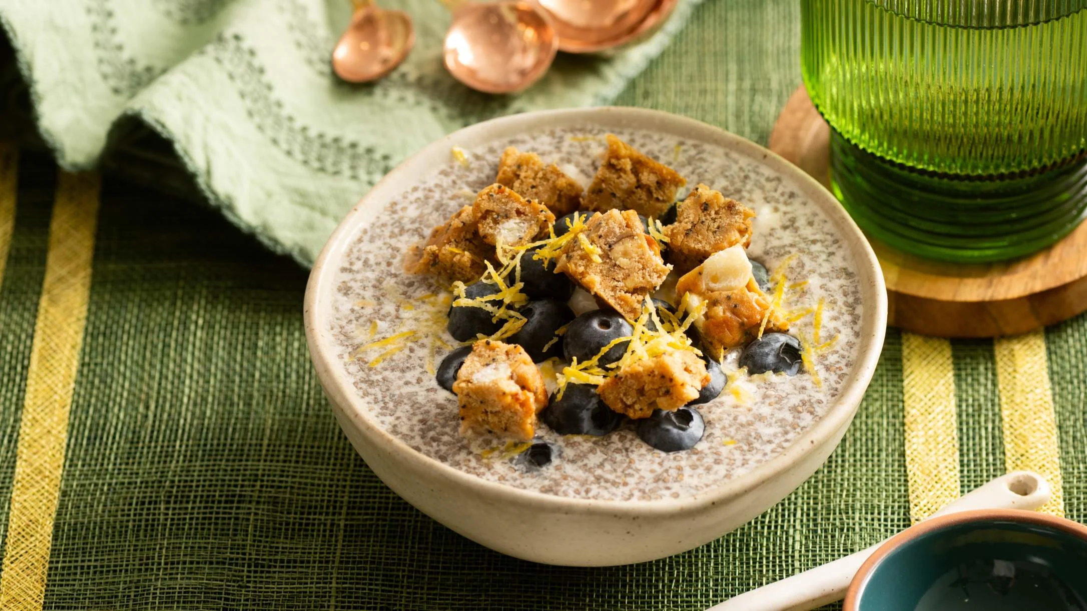 Creamy chia pudding topped with blueberries, Atkins protein bar, and lemon zest in a ceramic bowl on green placemat.