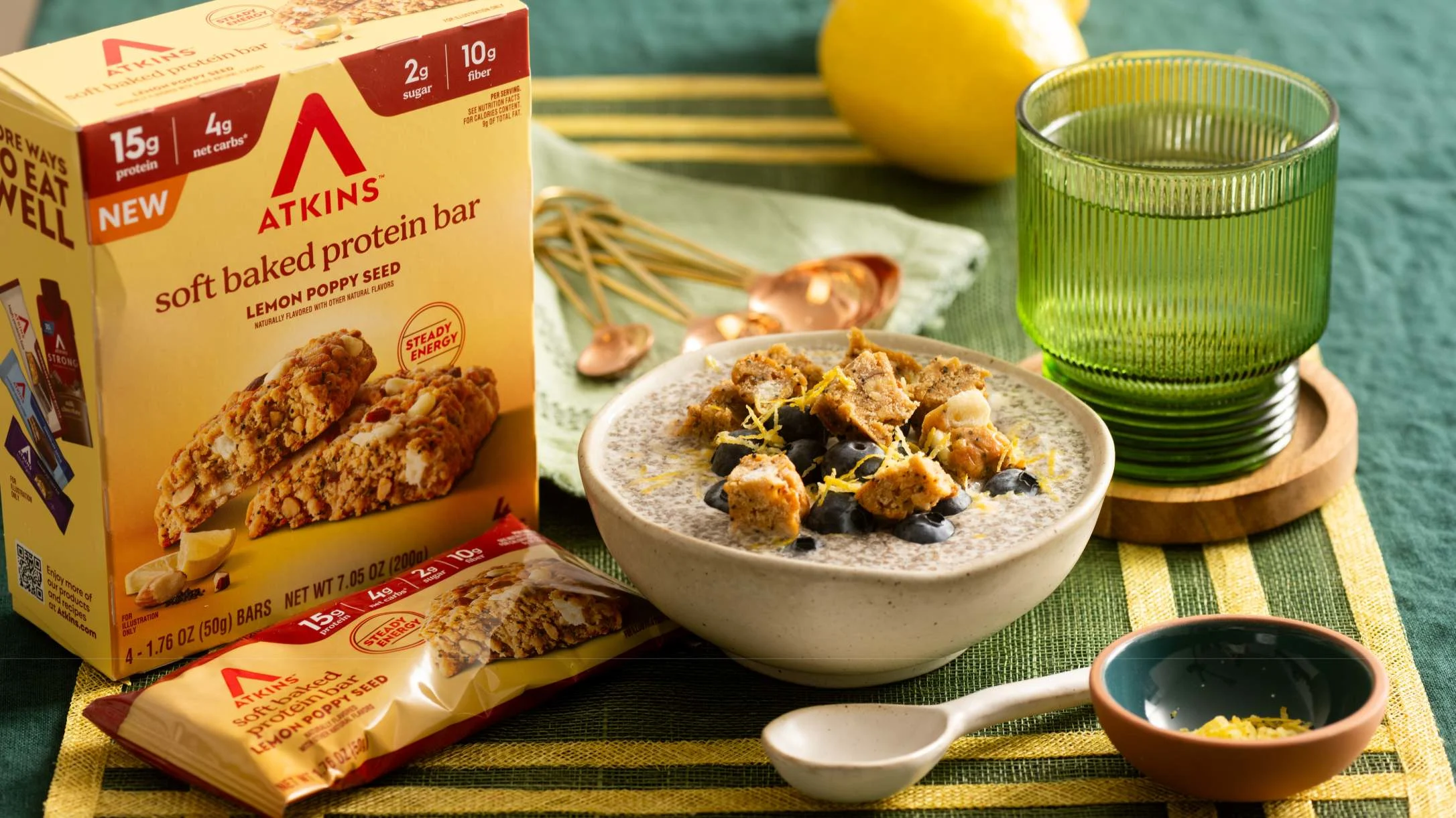 Atkins Lemon Poppy Seed protein bar box beside a bowl of chia pudding topped with bar pieces, blueberries, and lemon zest.