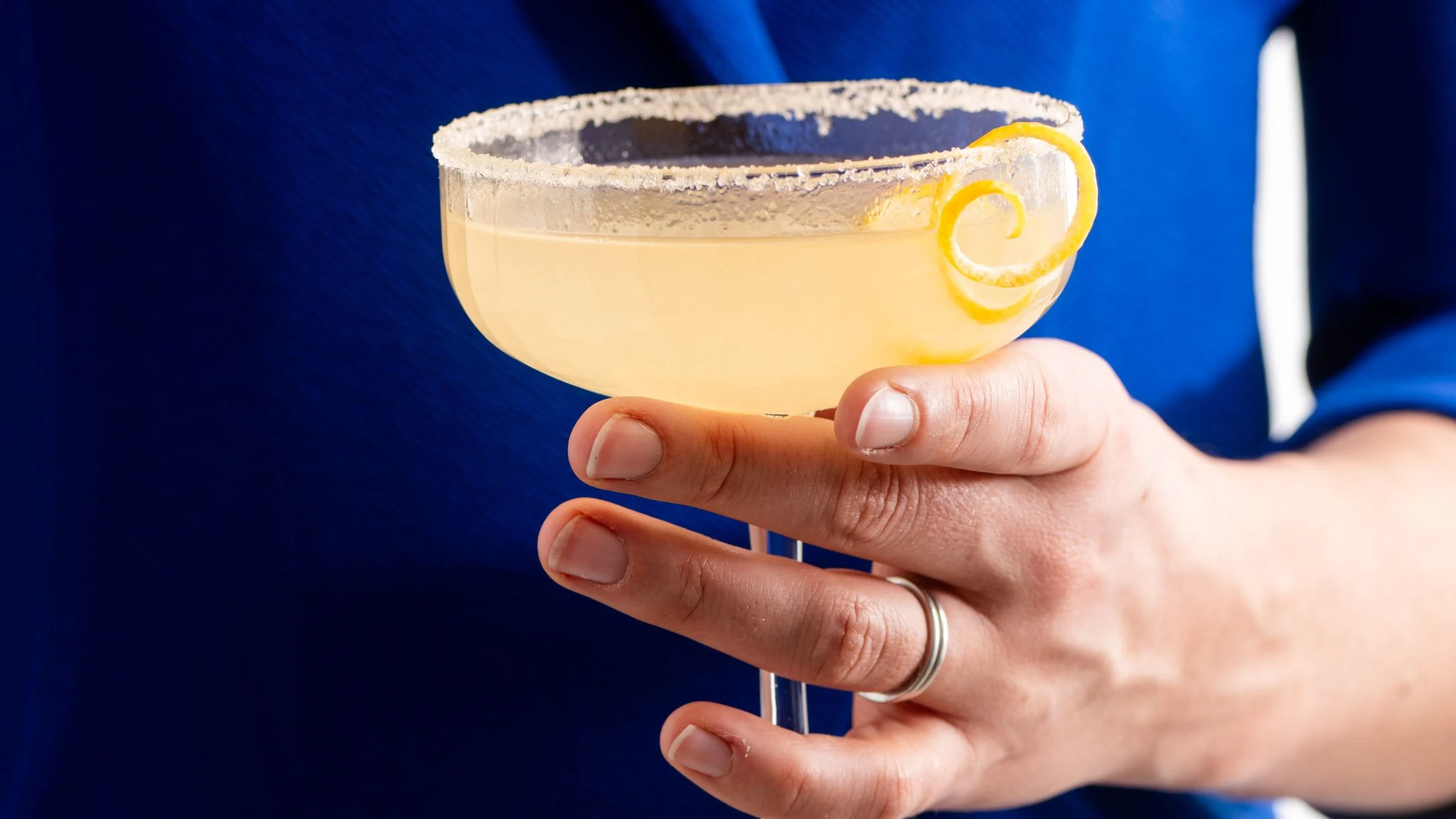 Hand holding a yellow cocktail with salt rim and lemon twist garnish against a vibrant blue background.