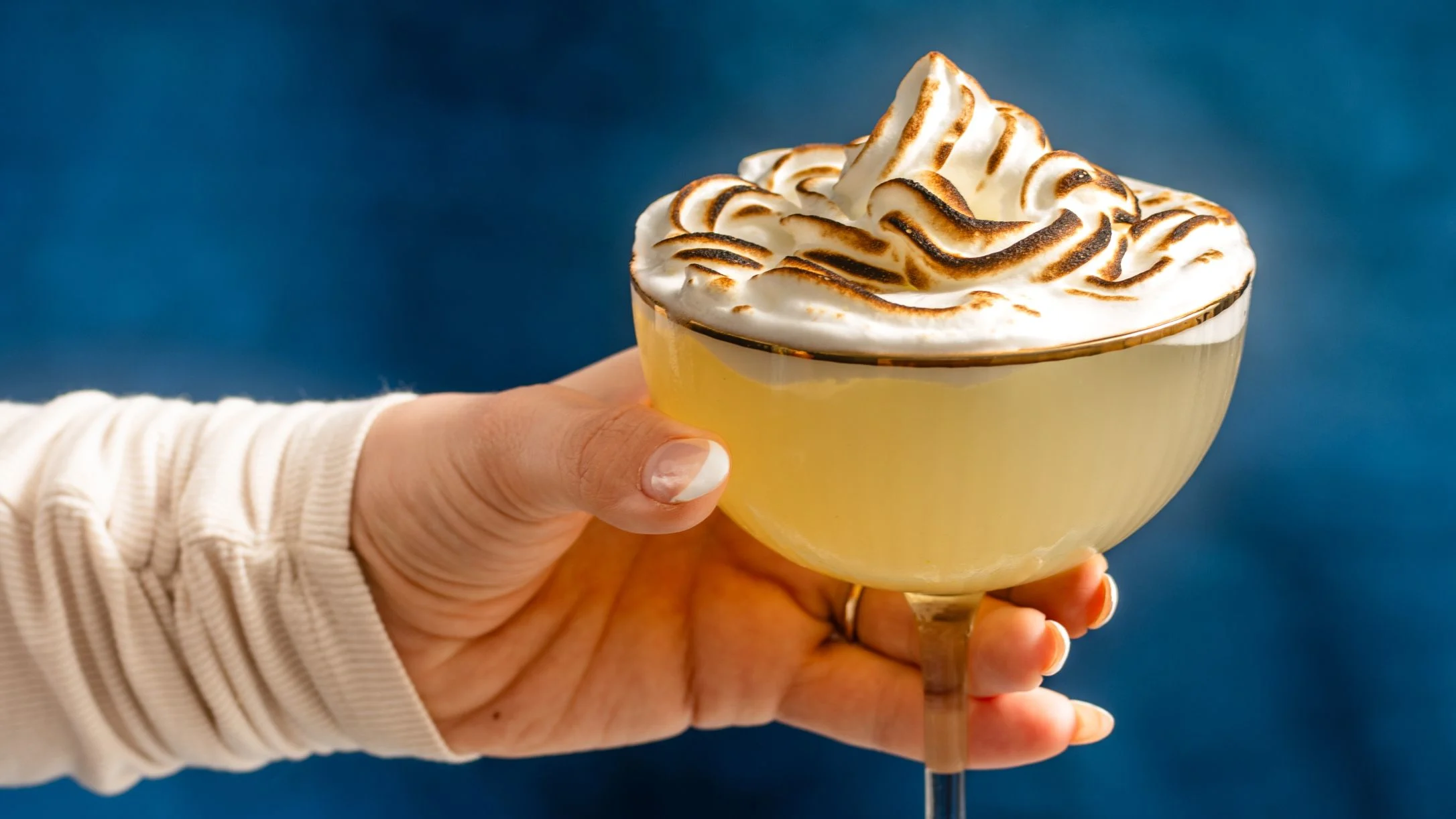 Hand holding a cocktail with toasted meringue topping in a coupe glass against a blue background.