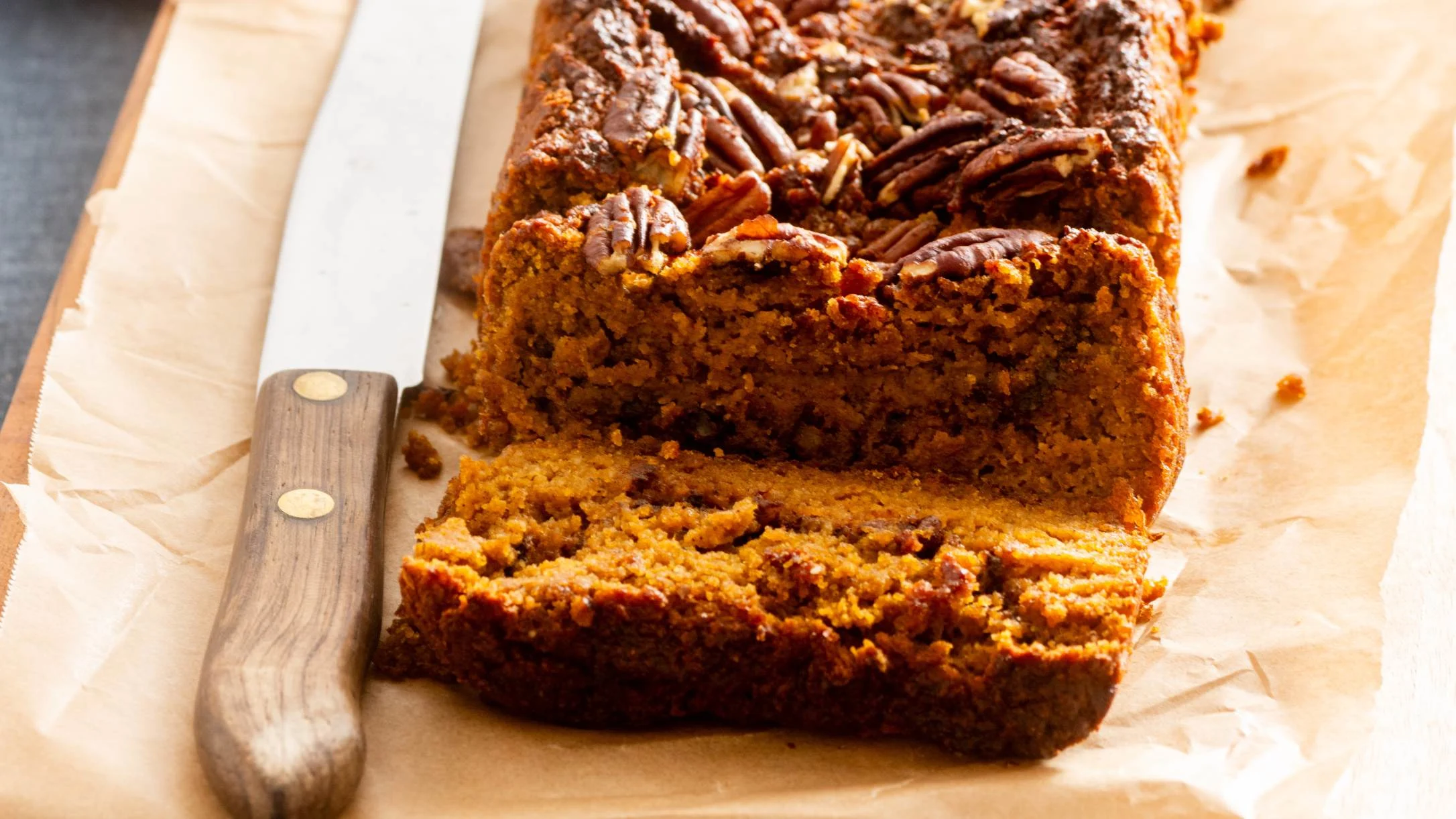 Freshly baked pumpkin bread with pecans on top, sliced to show moist interior, on parchment paper with knife.