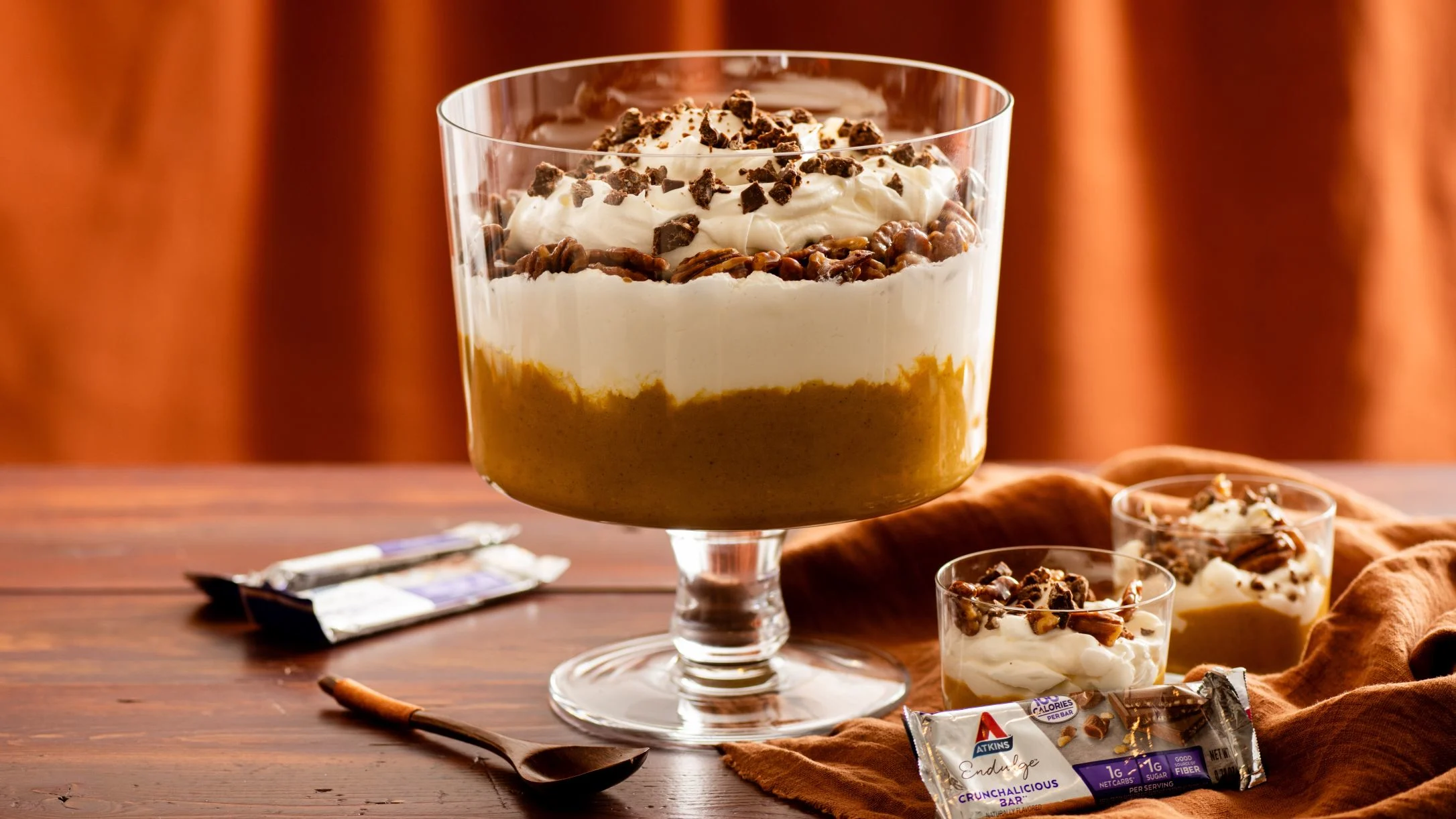 Layered pumpkin trifle dessert in glass dish with whipped cream and chocolate pieces, served with Atkins snack bars.
