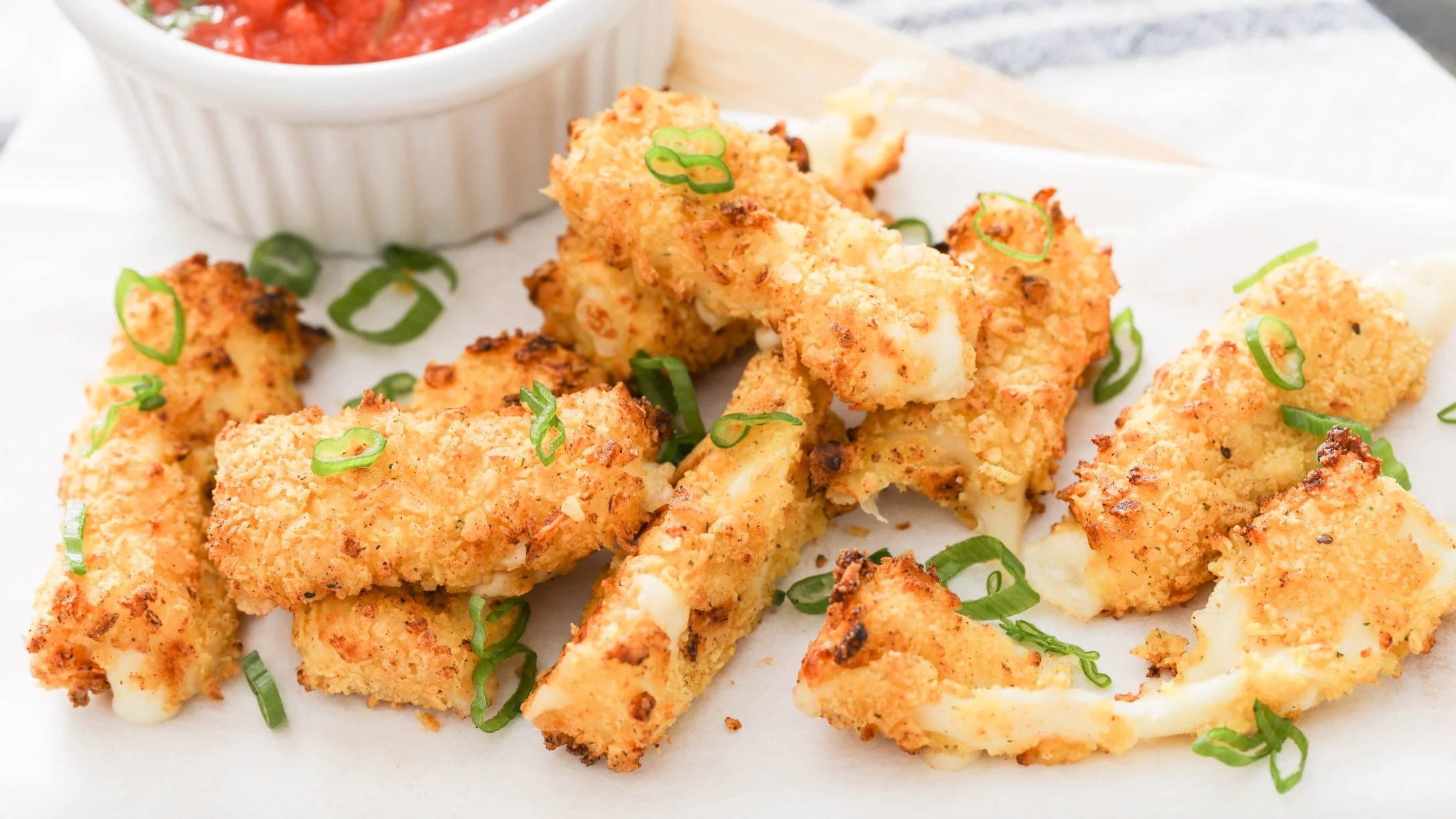 Crispy breaded cheese bites garnished with green onions, served with a red dipping sauce in a white bowl.