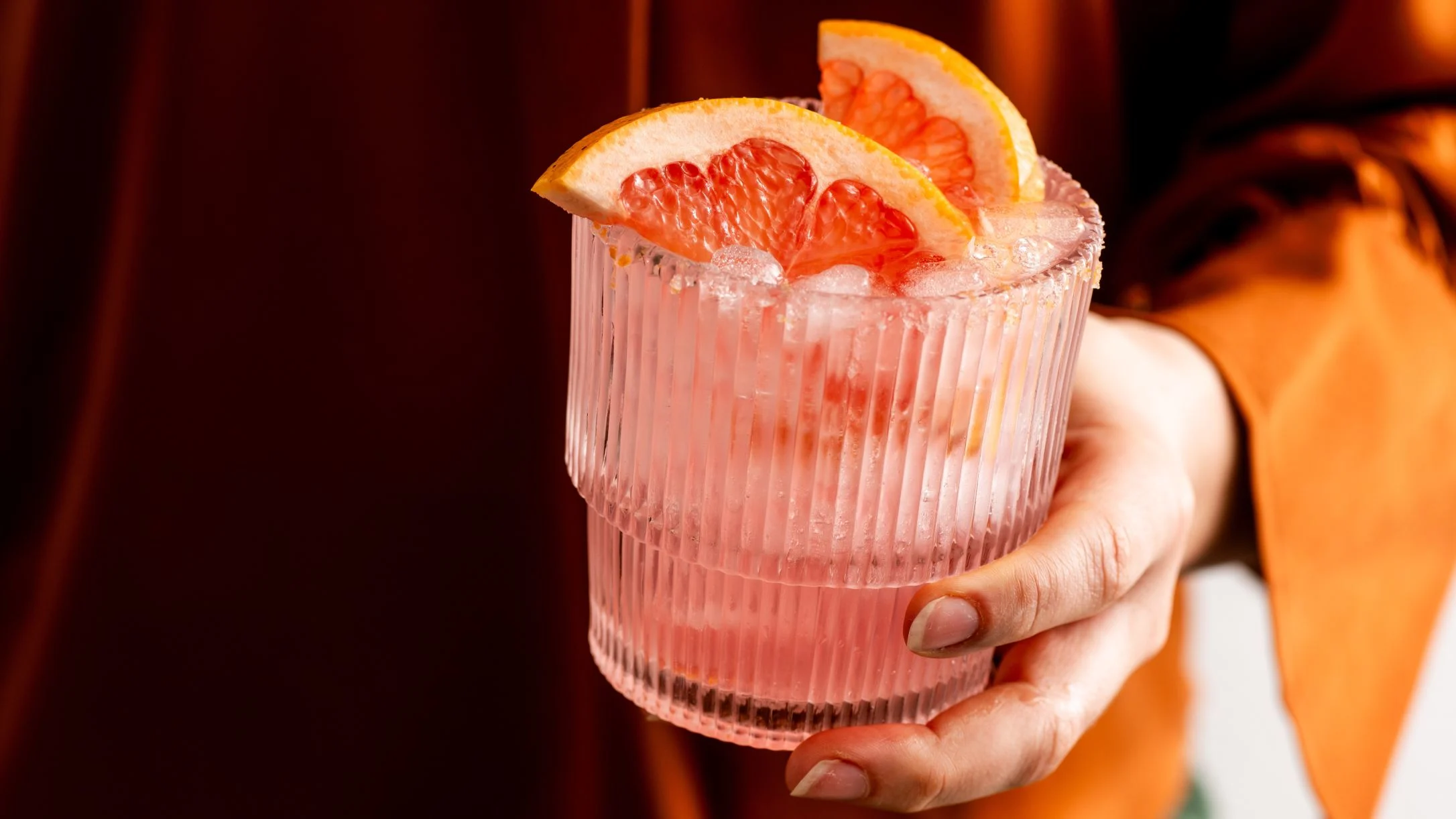 Hand holding a textured glass with pink cocktail garnished with grapefruit slices and ice in warm lighting.