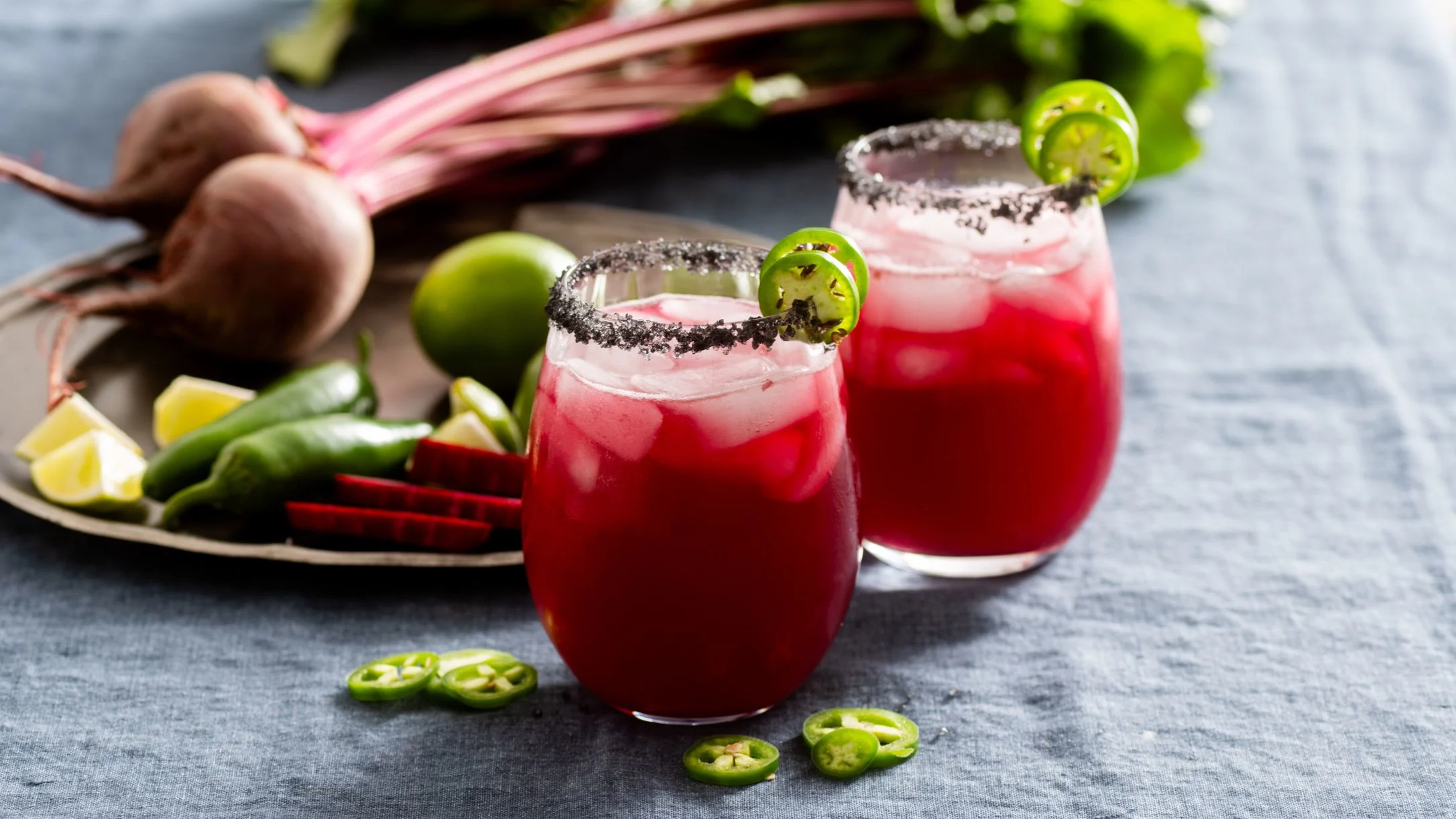 Two vibrant red beet cocktails with salt rims and lime garnish, served with fresh beets, limes, and jalapeño slices.
