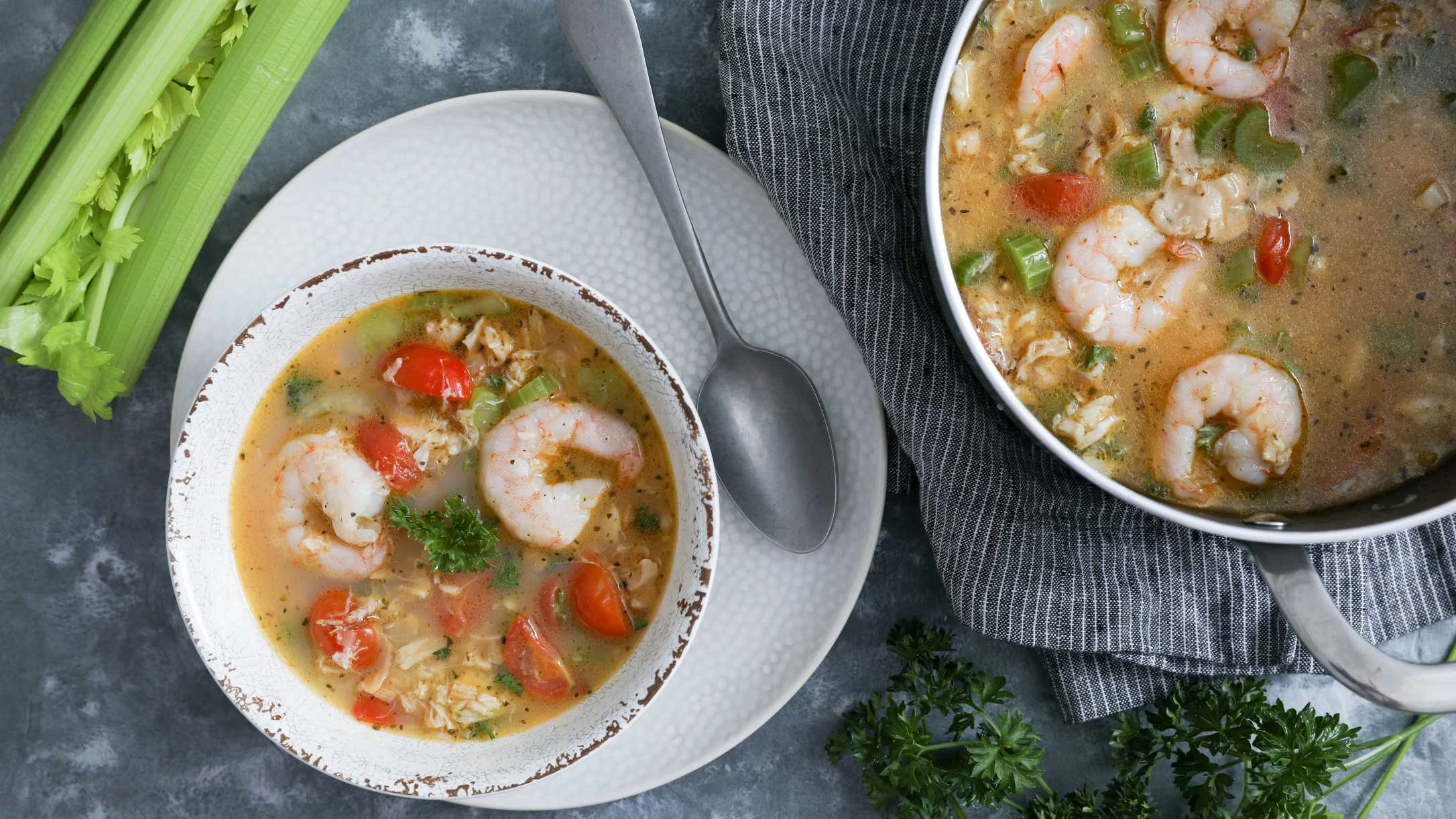 Shrimp seafood chowder in white bowl and pot with tomatoes, and vegetables, garnished with parsley and celery stalks.