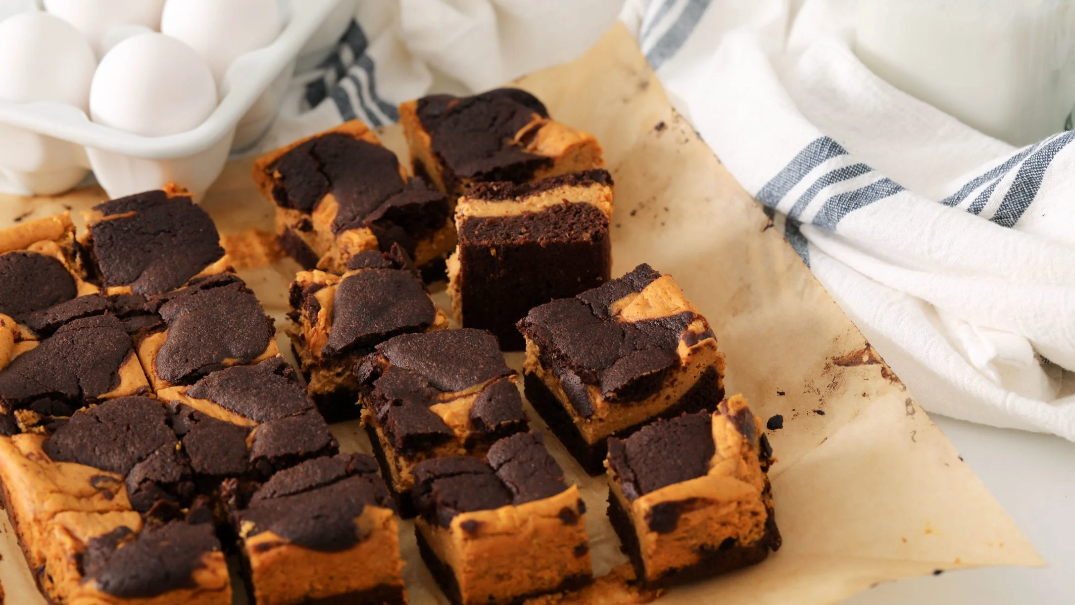 Chocolate pumpkin brownies cut into squares on parchment paper with egg carton and striped kitchen towel nearby.