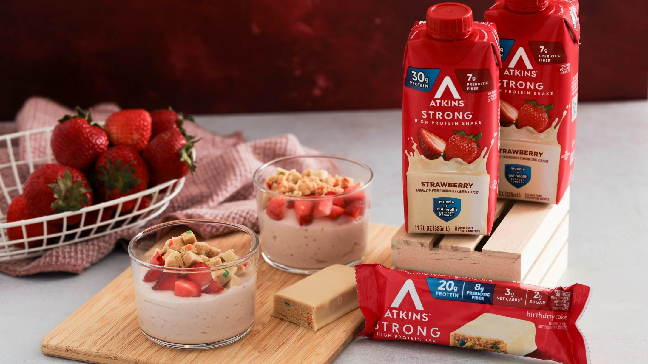 Atkins Strong protein shakes and bar with strawberry pudding parfaits and fresh strawberries on wooden board.