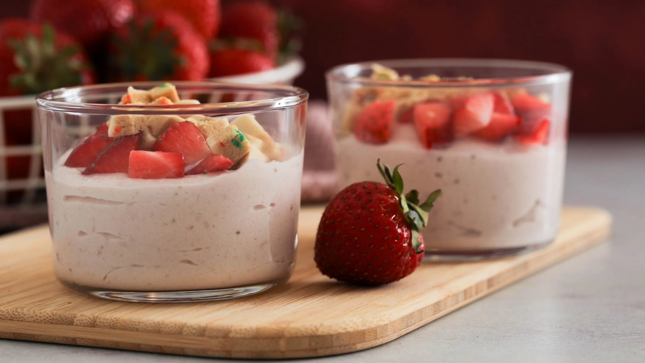 Two glass cups of creamy dessert topped with fresh strawberries on a wooden board, with a whole strawberry beside them.
