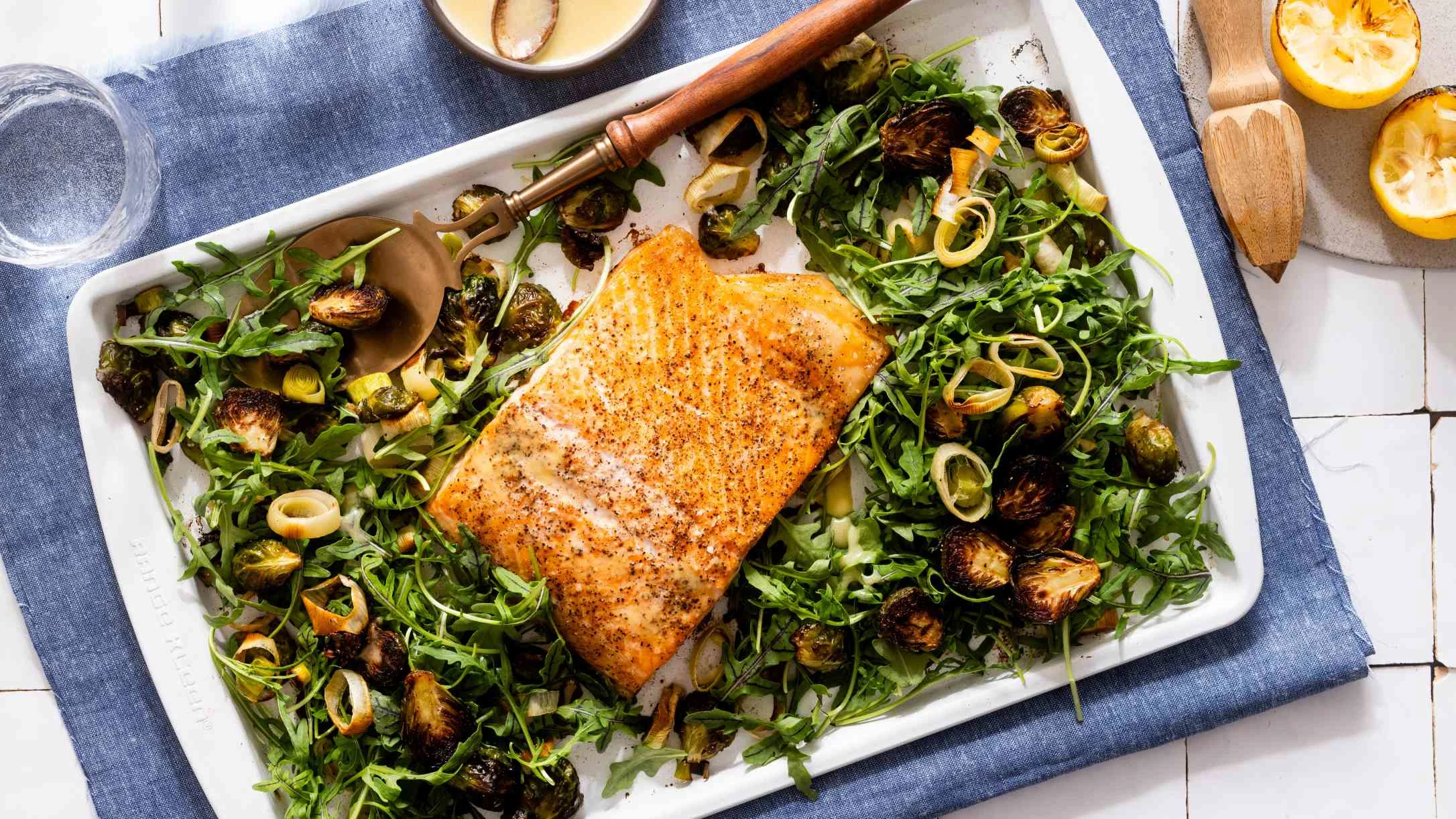 Roasted salmon fillet on white platter with arugula and roasted Brussels sprouts, served on blue cloth napkin.