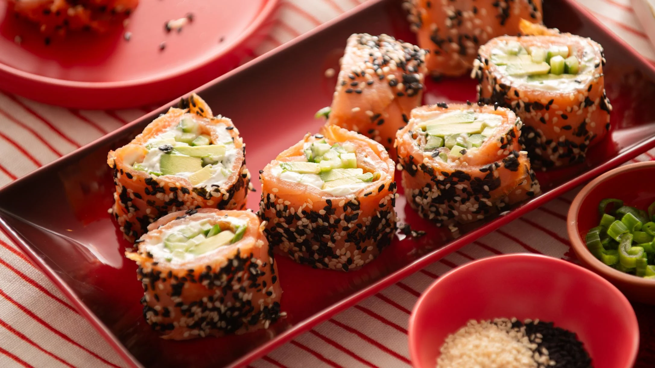 Salmon sushi rolls coated with sesame seeds, filled with cream cheese and avocado, served on a red rectangular plate.