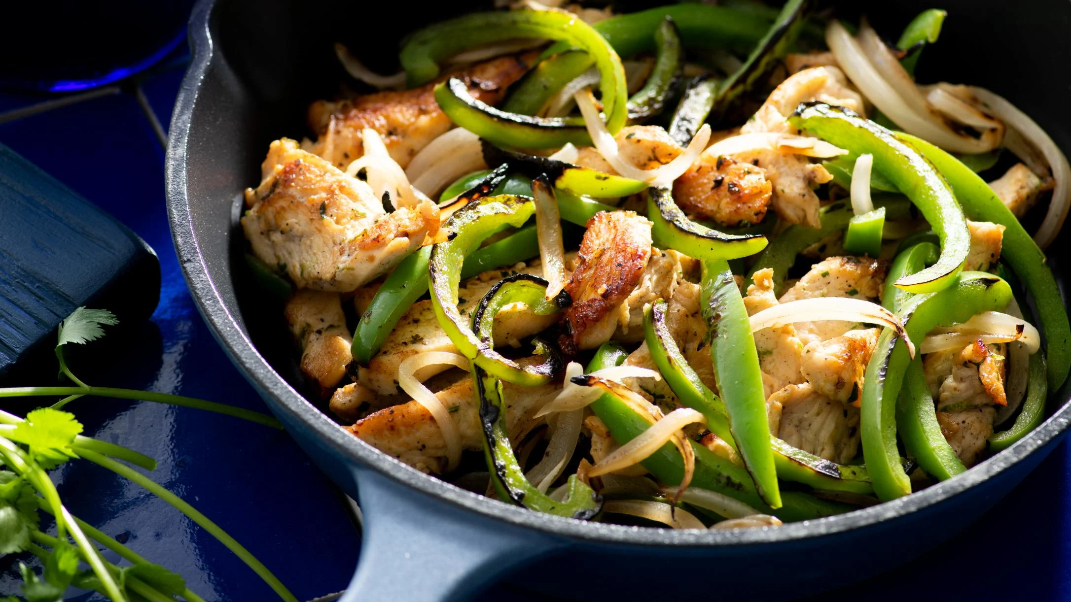 Sizzling chicken fajitas with green bell peppers and onions in a cast iron skillet against a dark blue background.