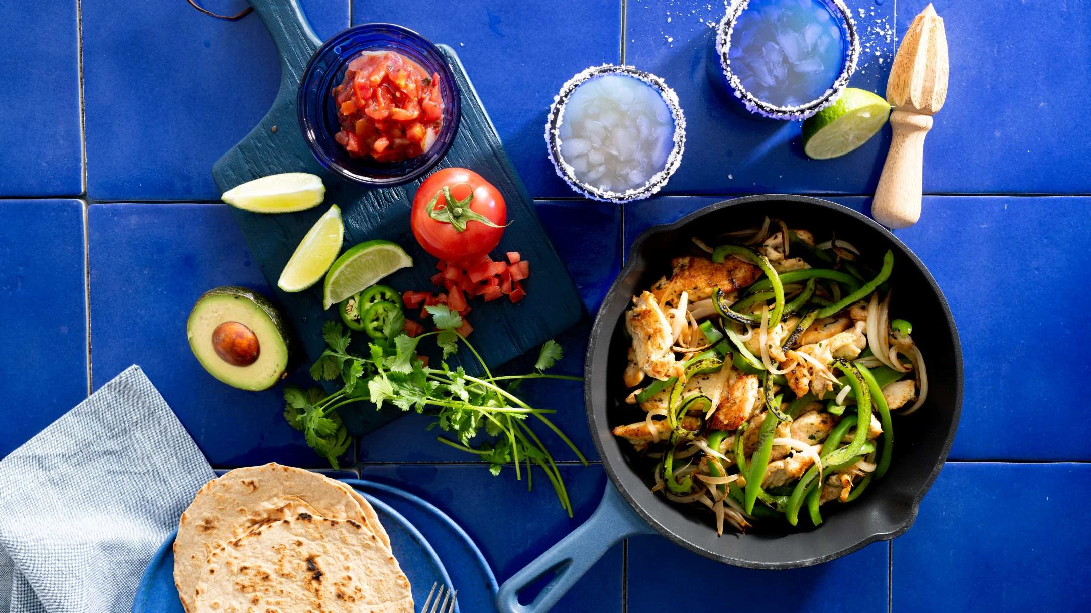 Mexican fajita ingredients on blue tile with skillet of chicken and peppers, salsa, avocado, lime, and tortillas.
